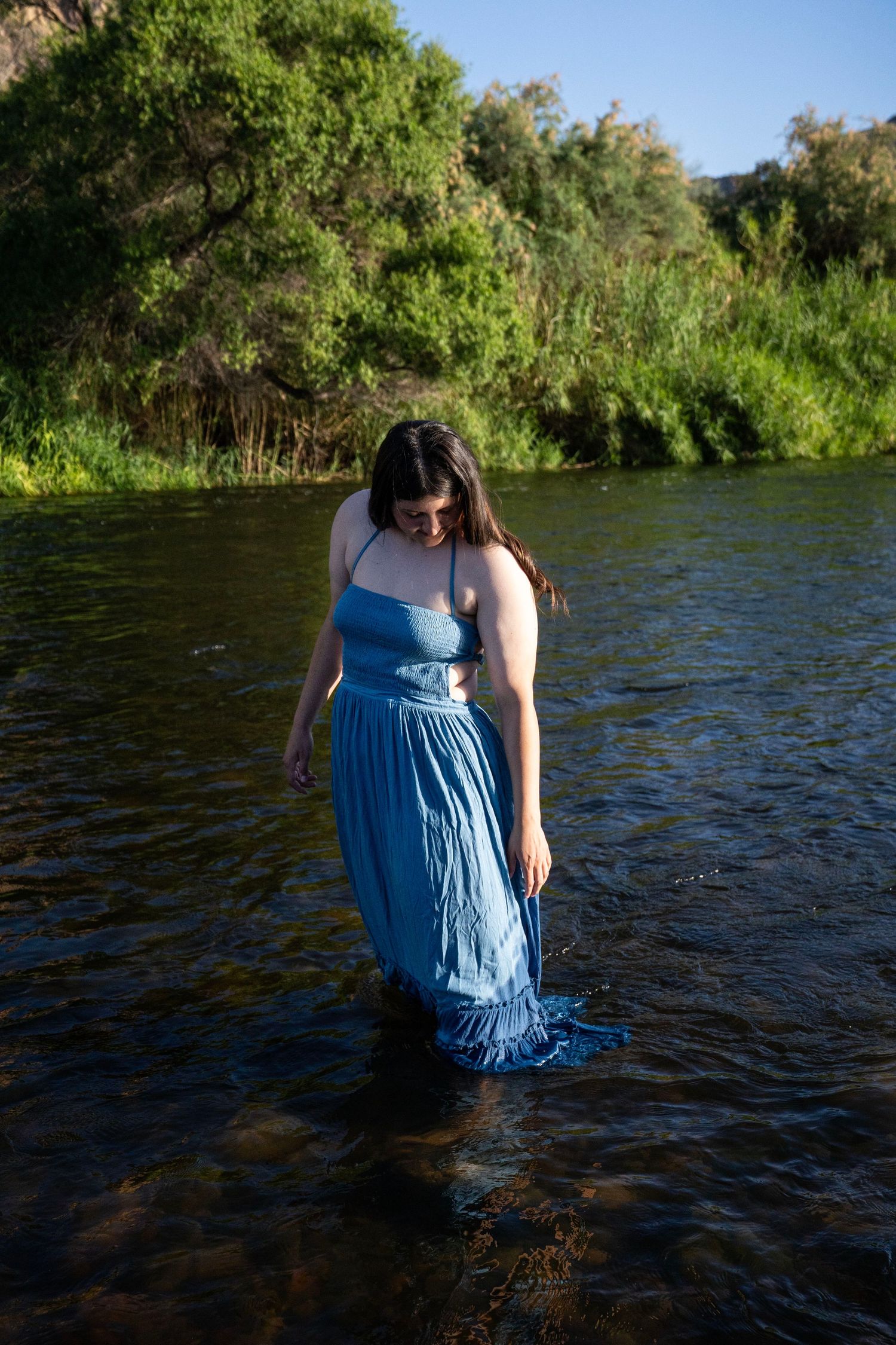 Woman in blue dress wading through a calm river surrounded by lush green trees and shrubs.