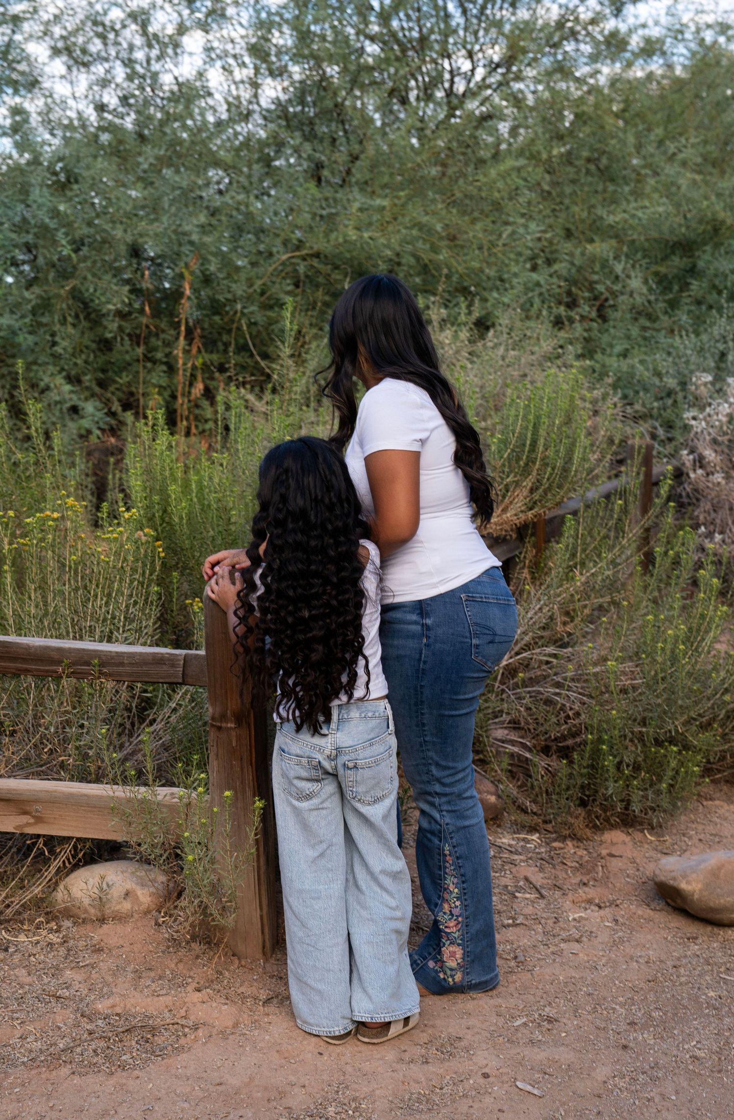 Woman and young girl with long curly hair looking at plants over a wooden fence outdoors.