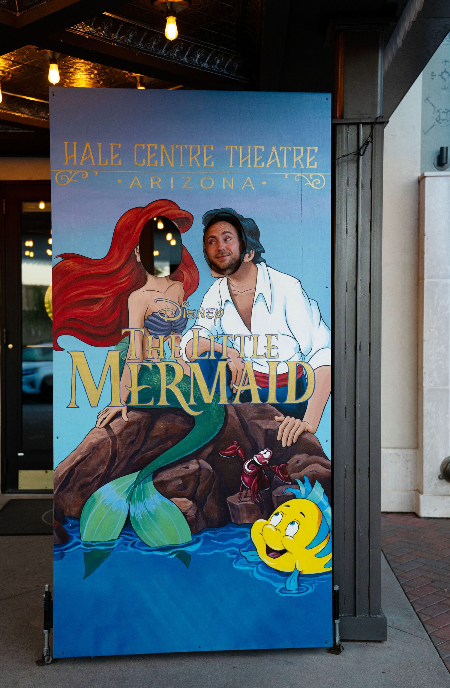 Man posing with The Little Mermaid promotional sign at Hale Centre Theatre Arizona.