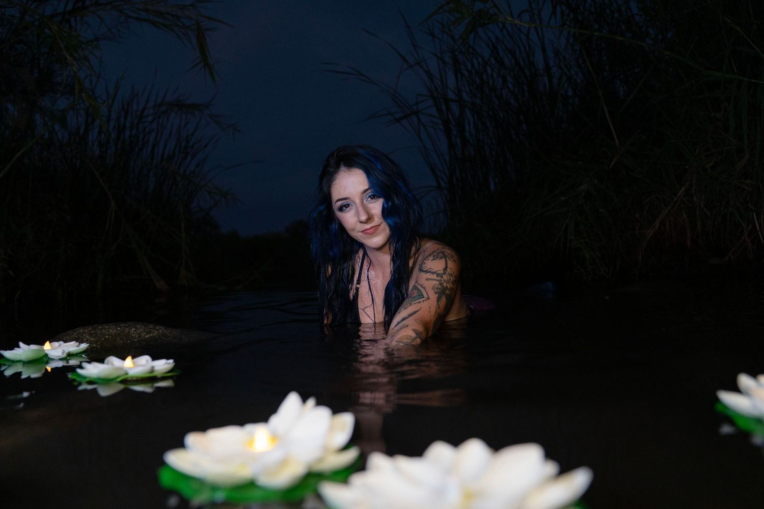 Tattooed woman emerging from dark water surrounded by white water lily flowers at night.