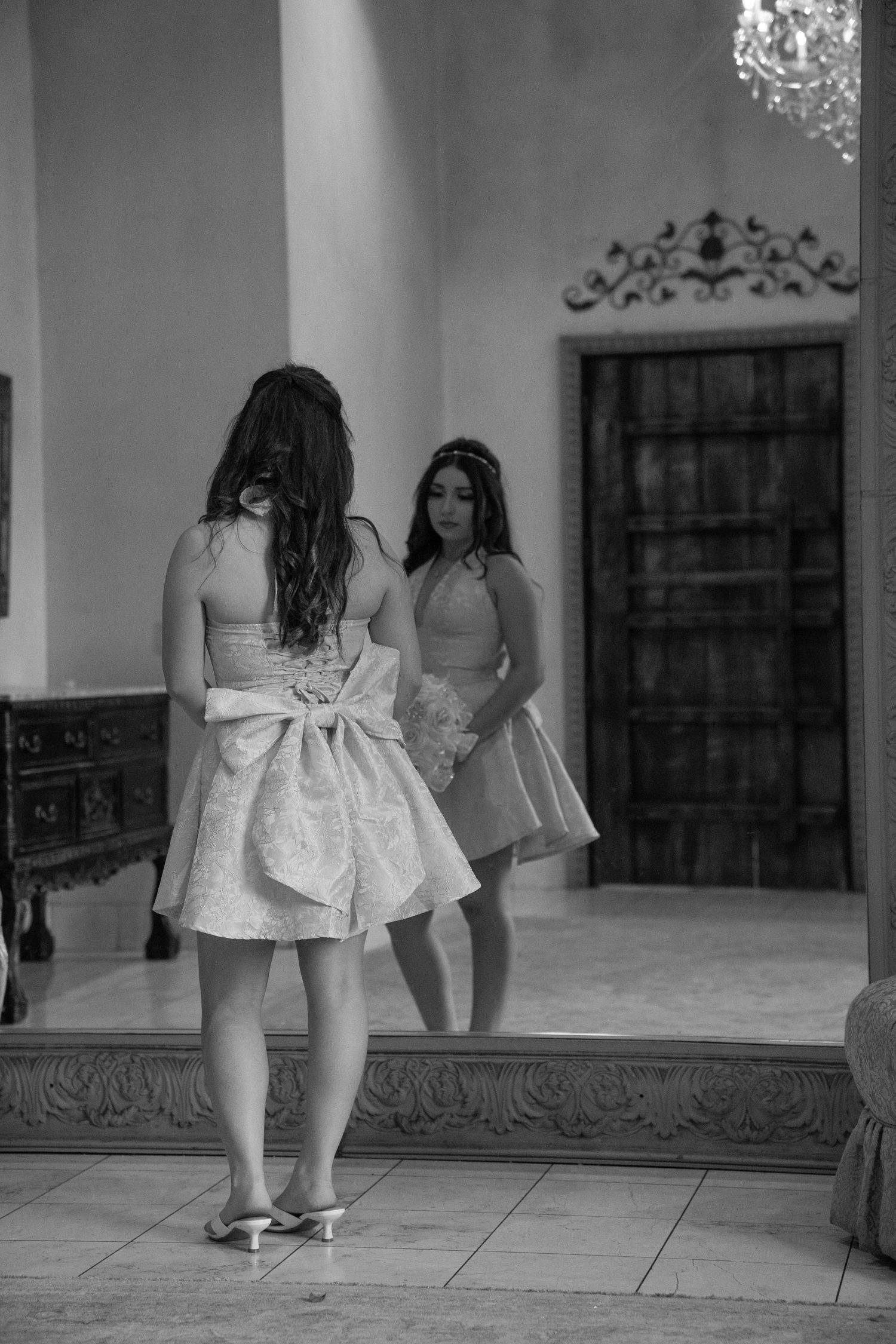 Black and white image of girl in pink dress standing before large elegant mirror.