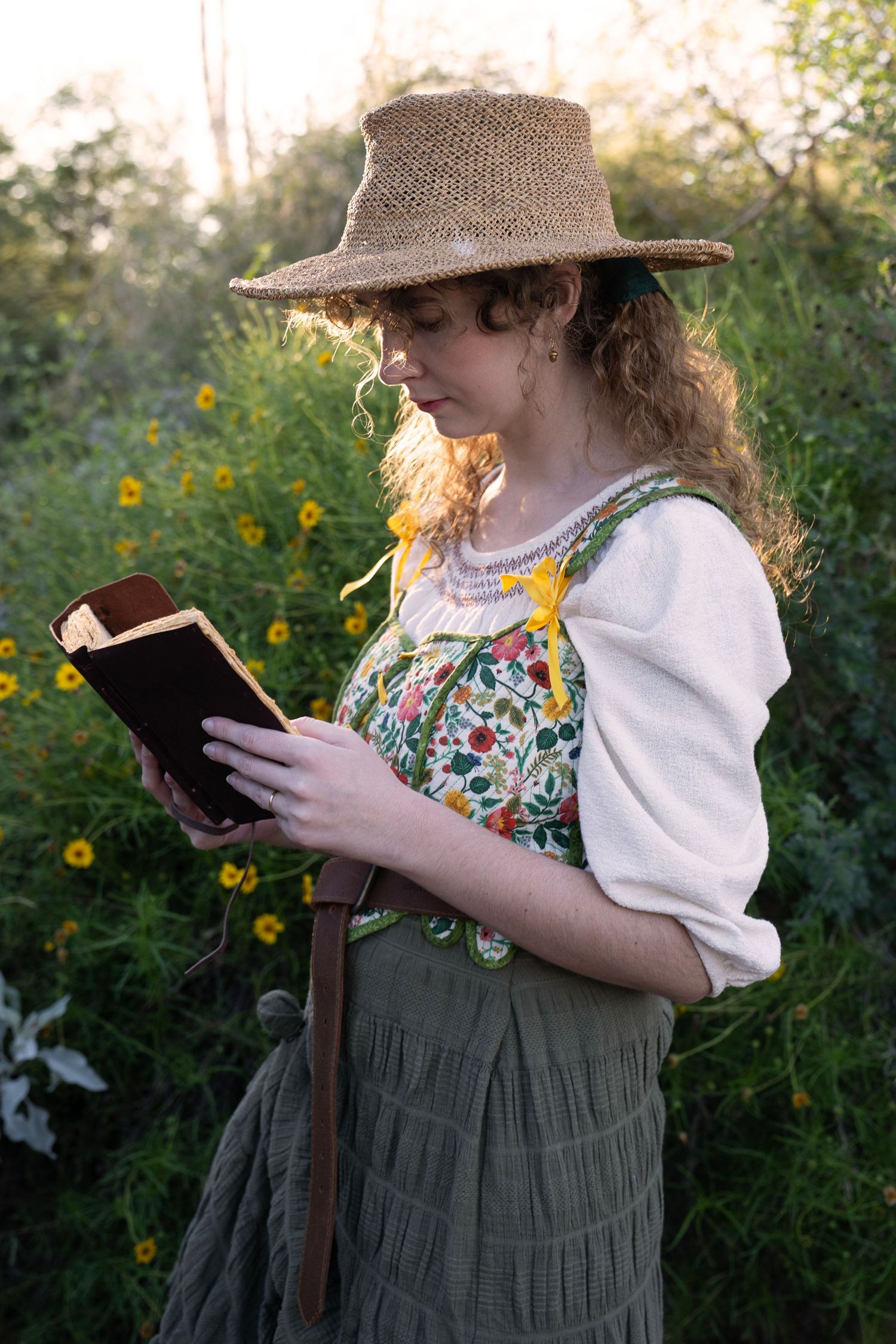 Woman in a straw hat and floral embroidered vest reads a book beside yellow wildflowers in soft golden light.