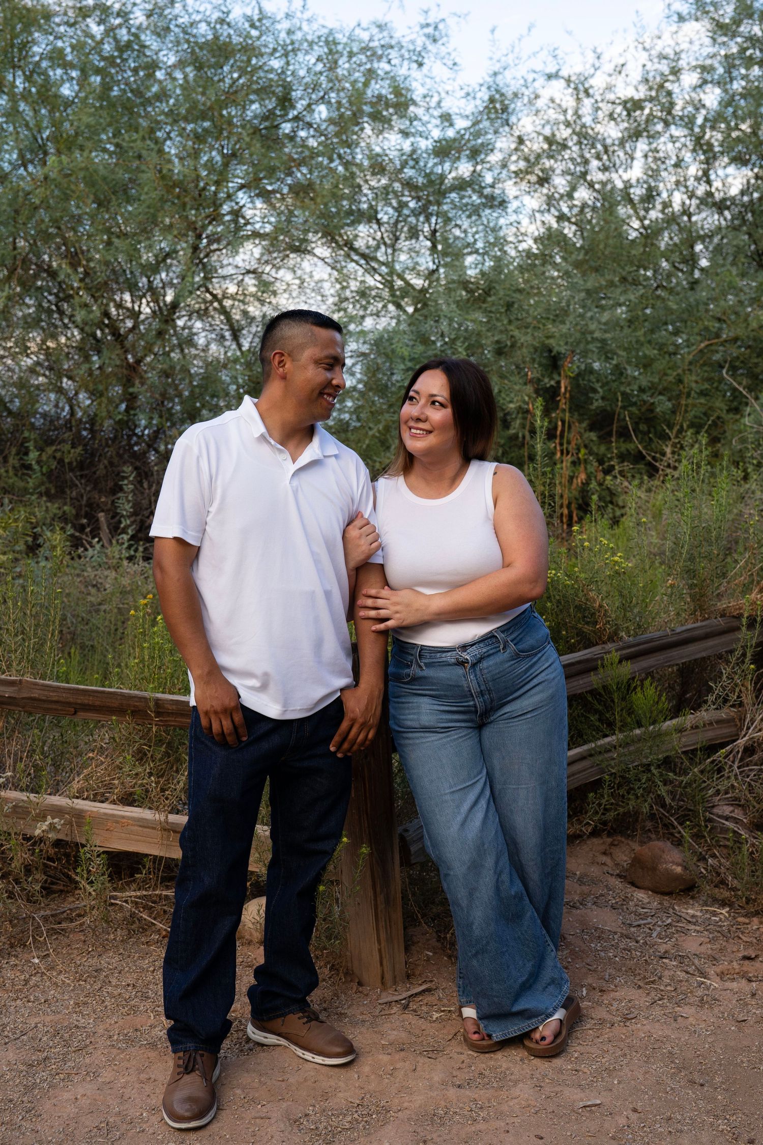 A couple in matching white tops and jeans smiles at each other while standing by a wooden fence in a lush outdoor setting.