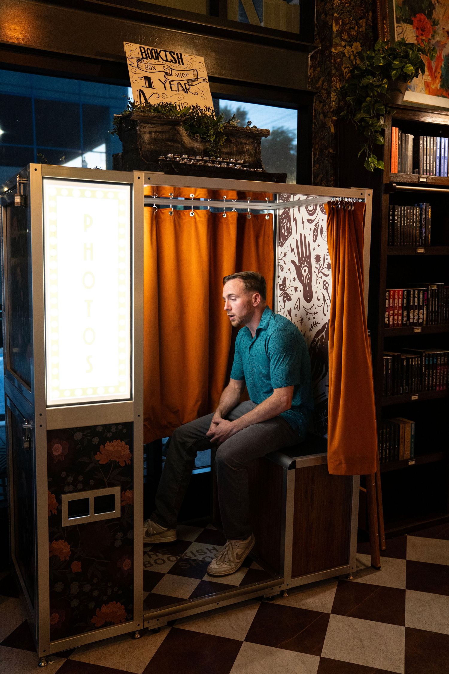 Man in teal polo shirt sitting inside vintage photo booth with orange curtains in bookstore.