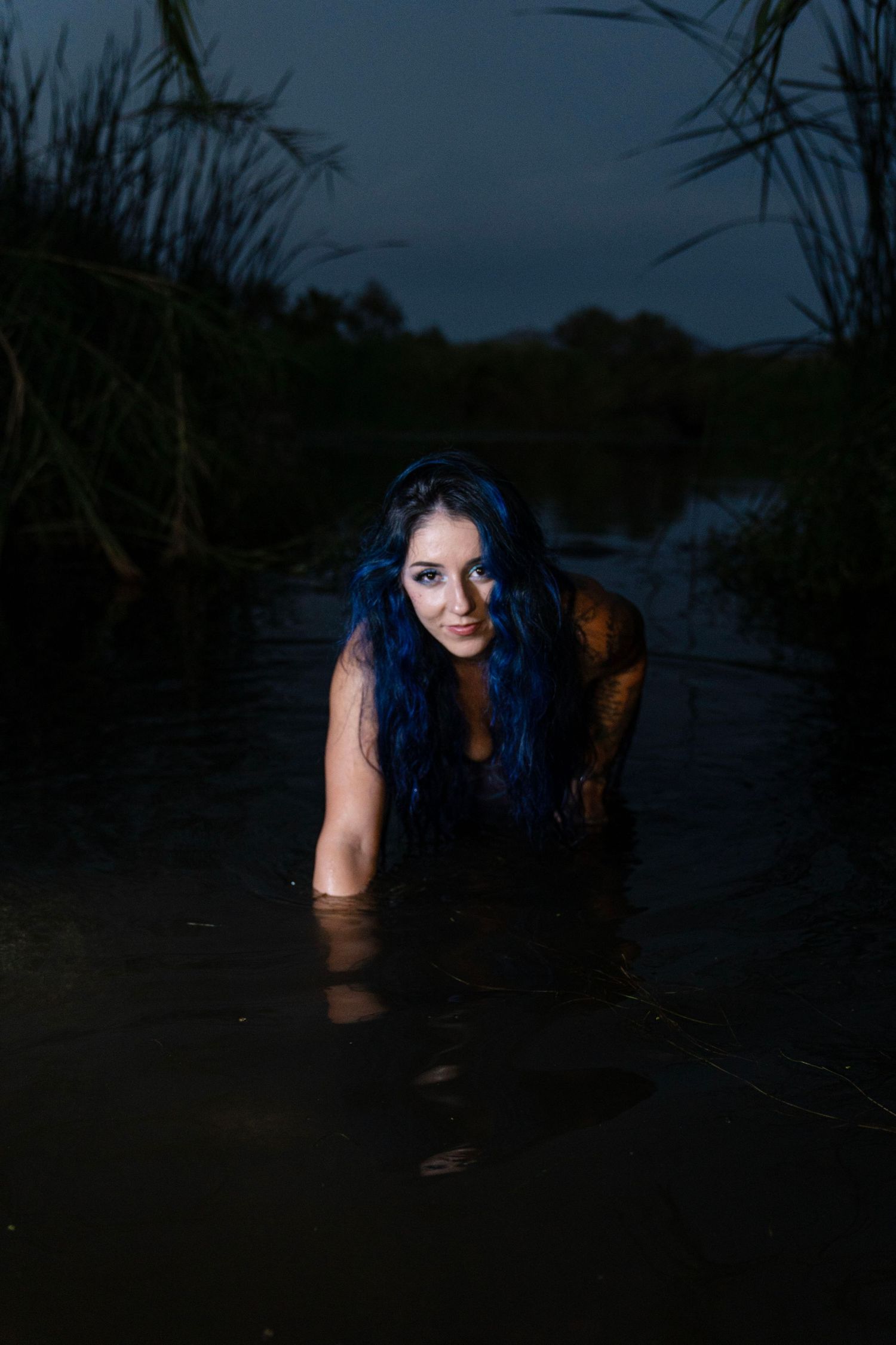 Blue-haired tattooed woman emerging from dark river water at night among tall reeds.