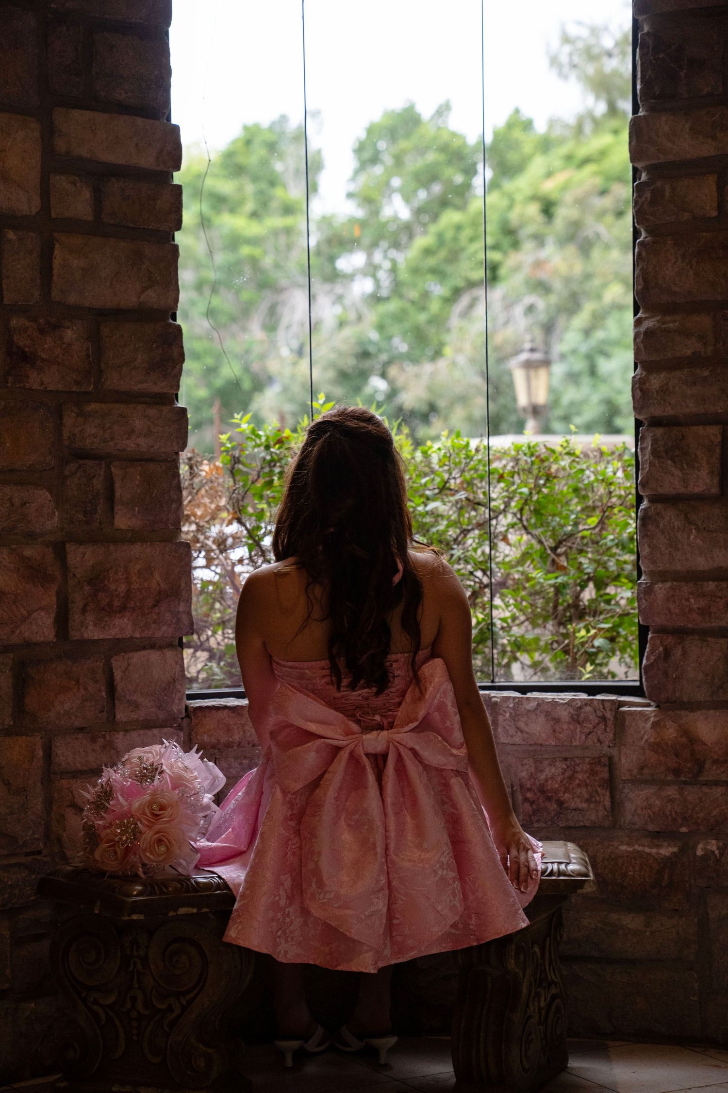 Back view of woman in pink bow dress standing before tall window holding floral bouquet.