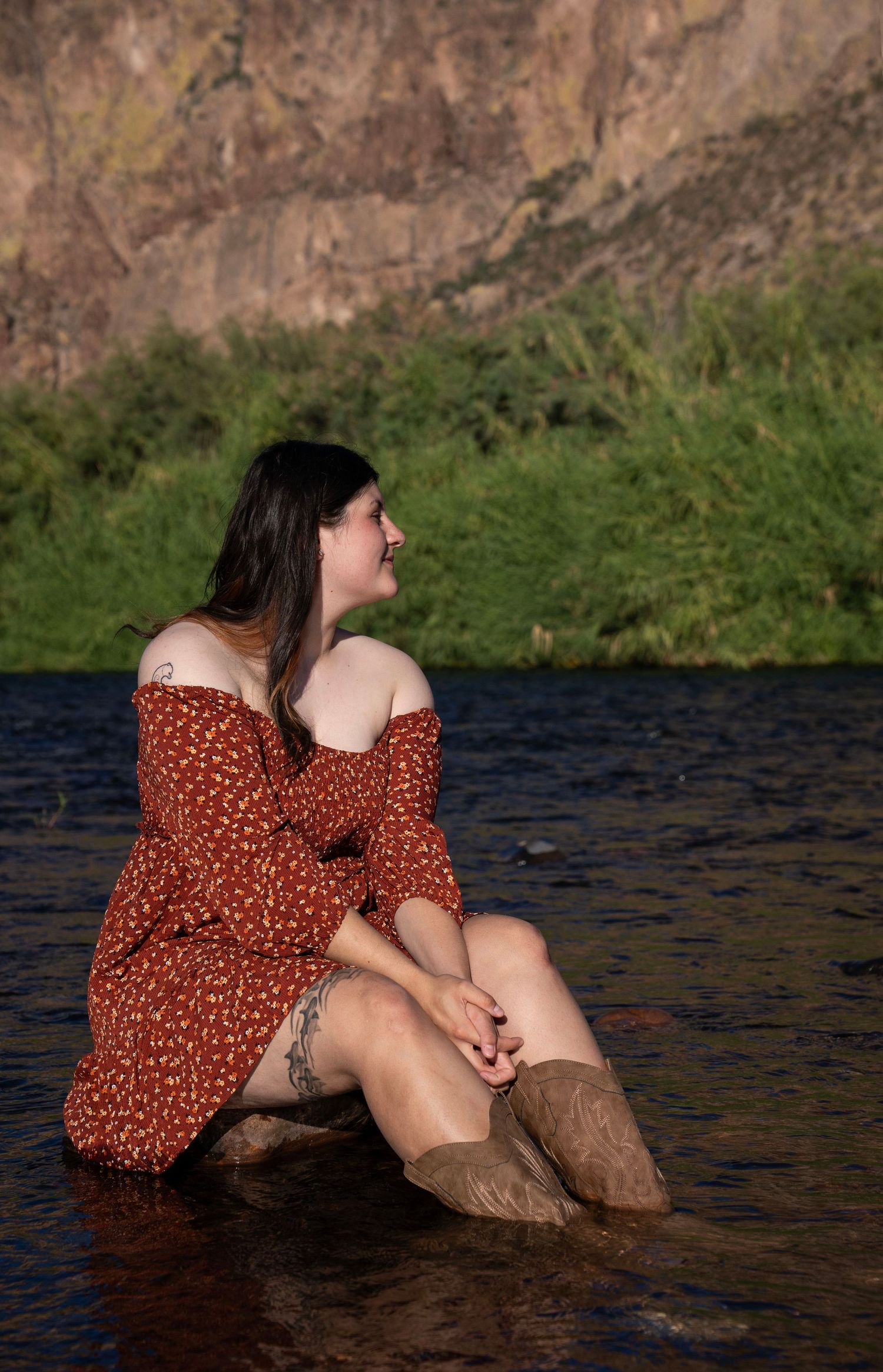 A smiling woman in a red floral off-shoulder dress sits in a shallow river wearing brown boots.