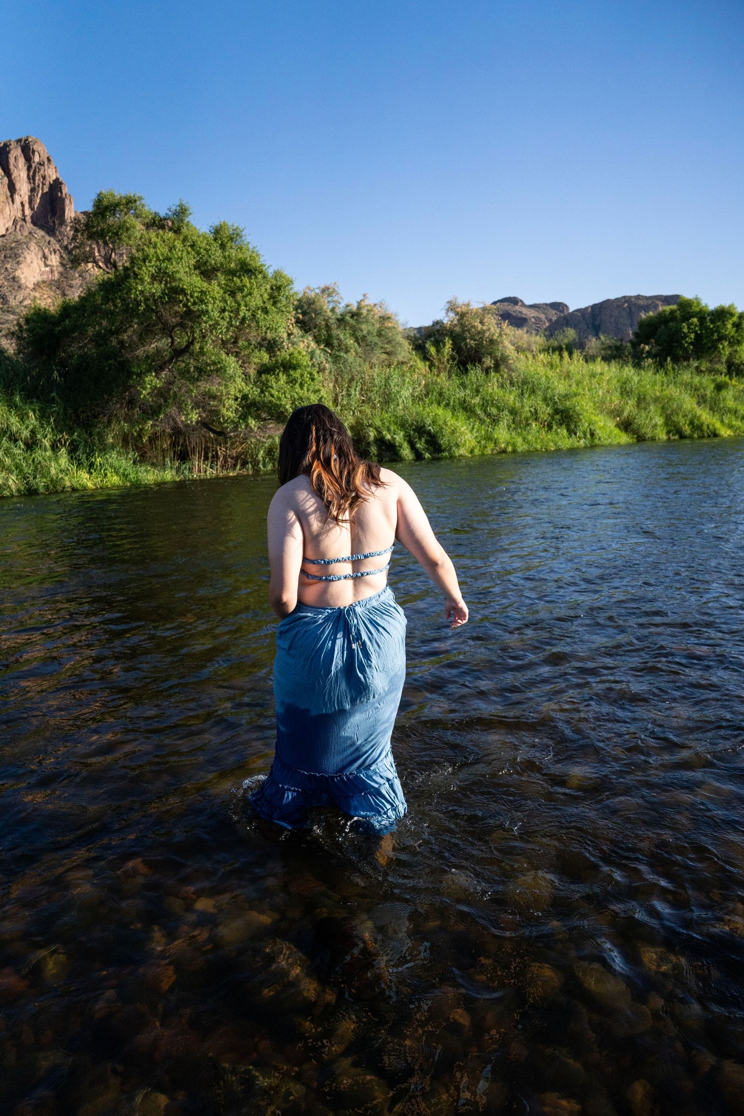 Woman wading in a scenic river wearing a blue sarong, surrounded by lush greenery and rocky cliffs.