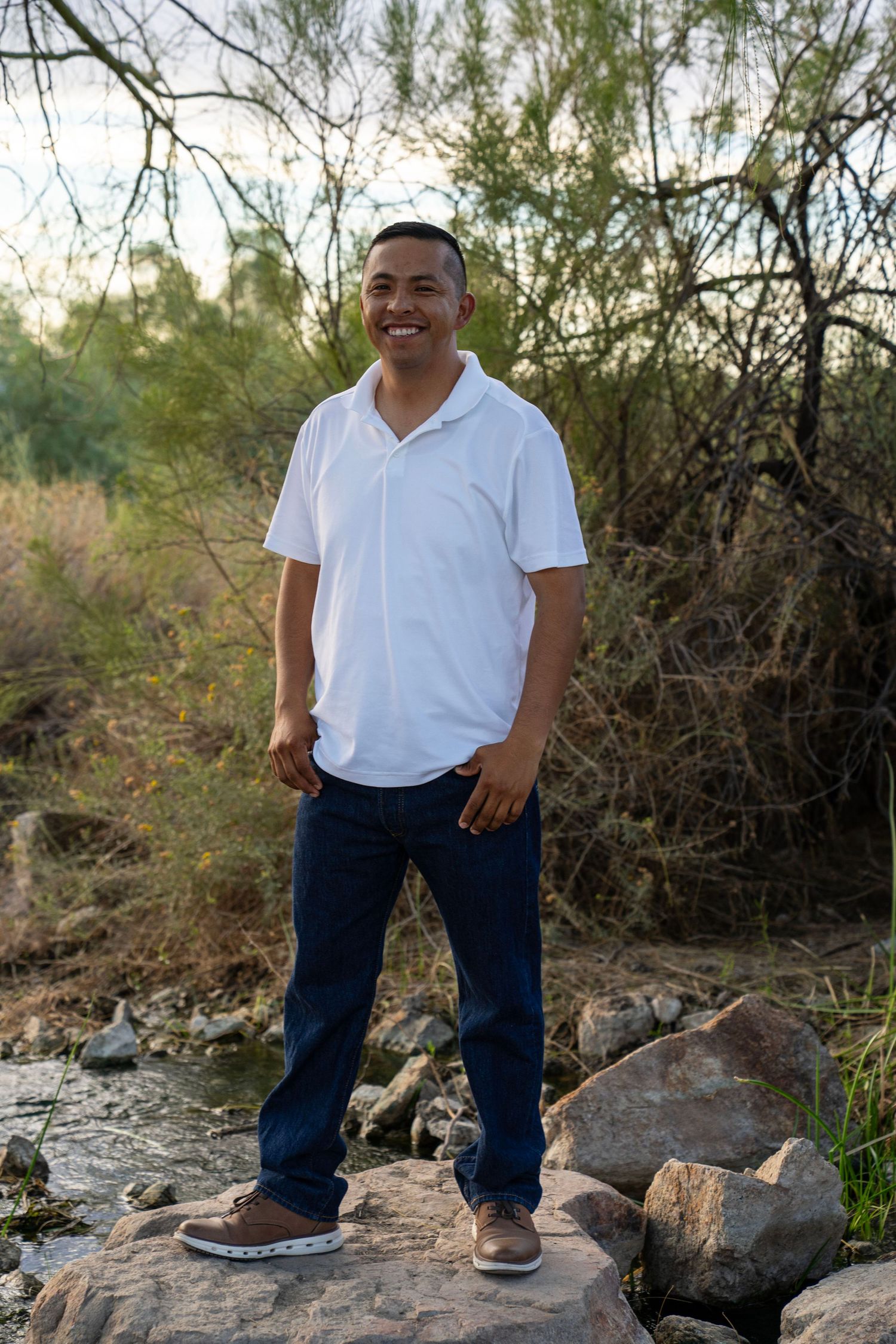 Happy man in white polo shirt standing on rocks near a stream surrounded by desert vegetation.