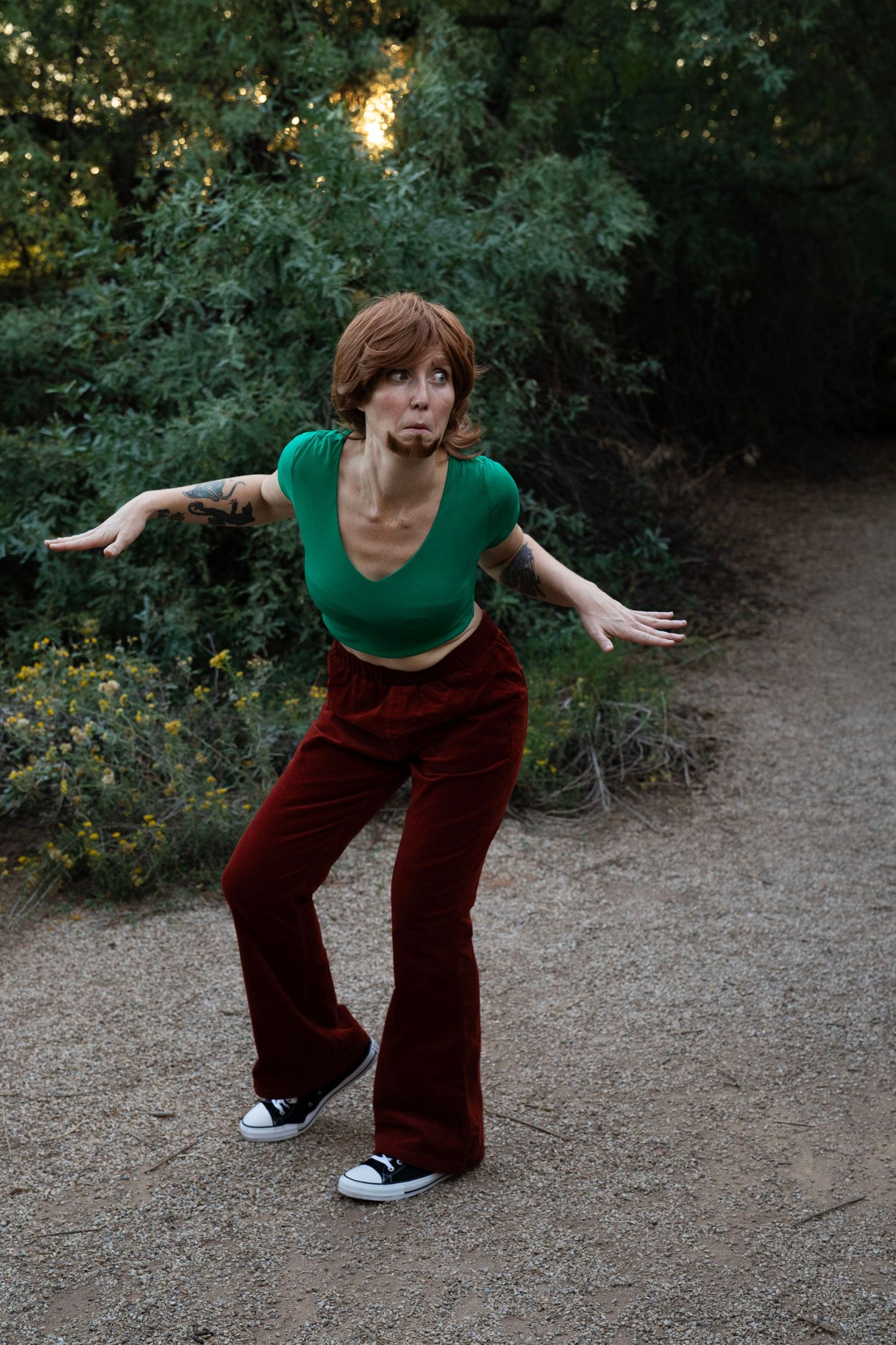 Person with short red hair in a green crop top, maroon flared pants and sneakers strikes a crouching pose on a gravel path by shrubs.