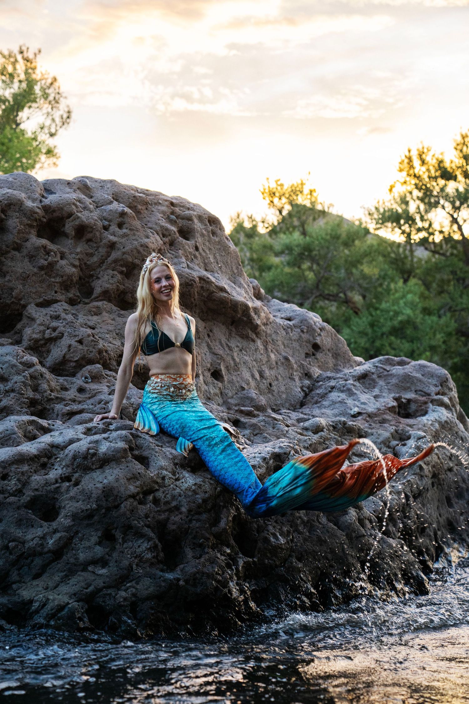 Smiling blonde woman in blue mermaid tail costume sitting on rocky shore at sunset. Mermaid photoshoot, fantasy photoshoot.