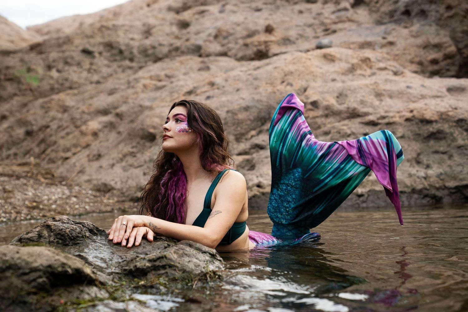A woman in a mermaid costume with purple and teal tail poses elegantly on rocks by water. Fantasy Photoshoot