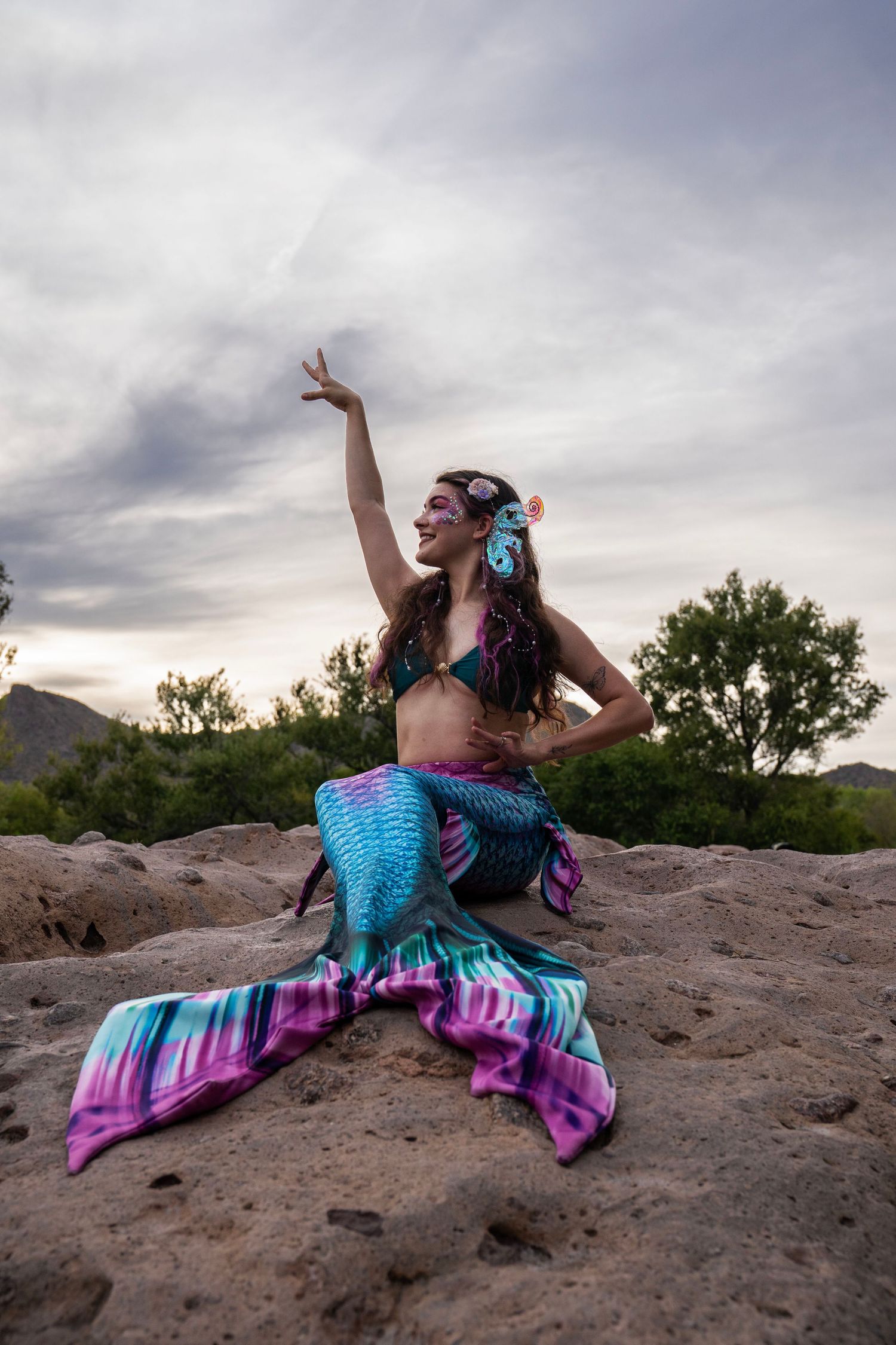 Woman in a colorful mermaid costume with teal and purple tail posing on rocks at sunset. Fantasy Photoshoot