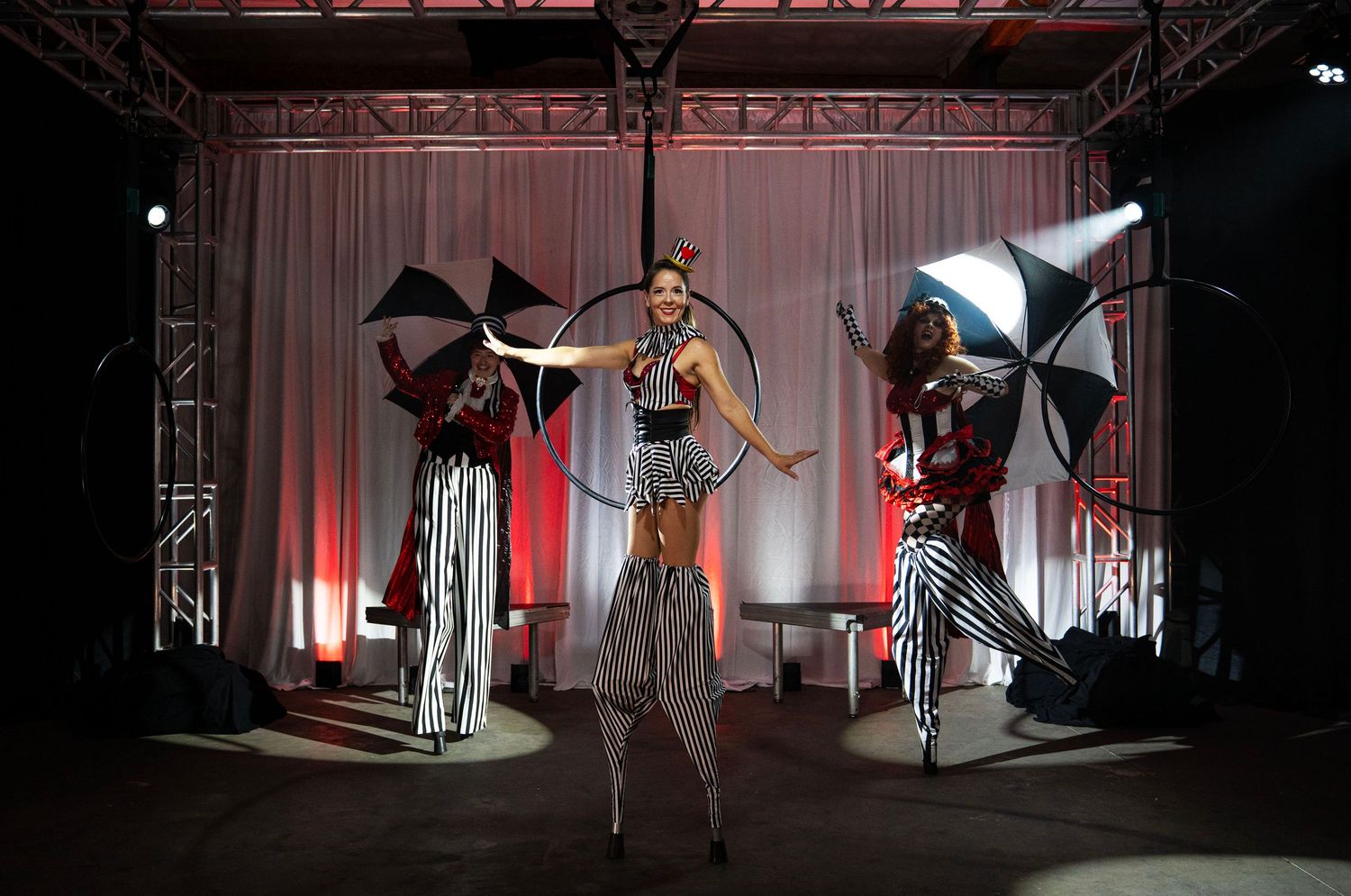 Three circus performers on stilts with hula hoops and umbrellas on red and white lit stage.