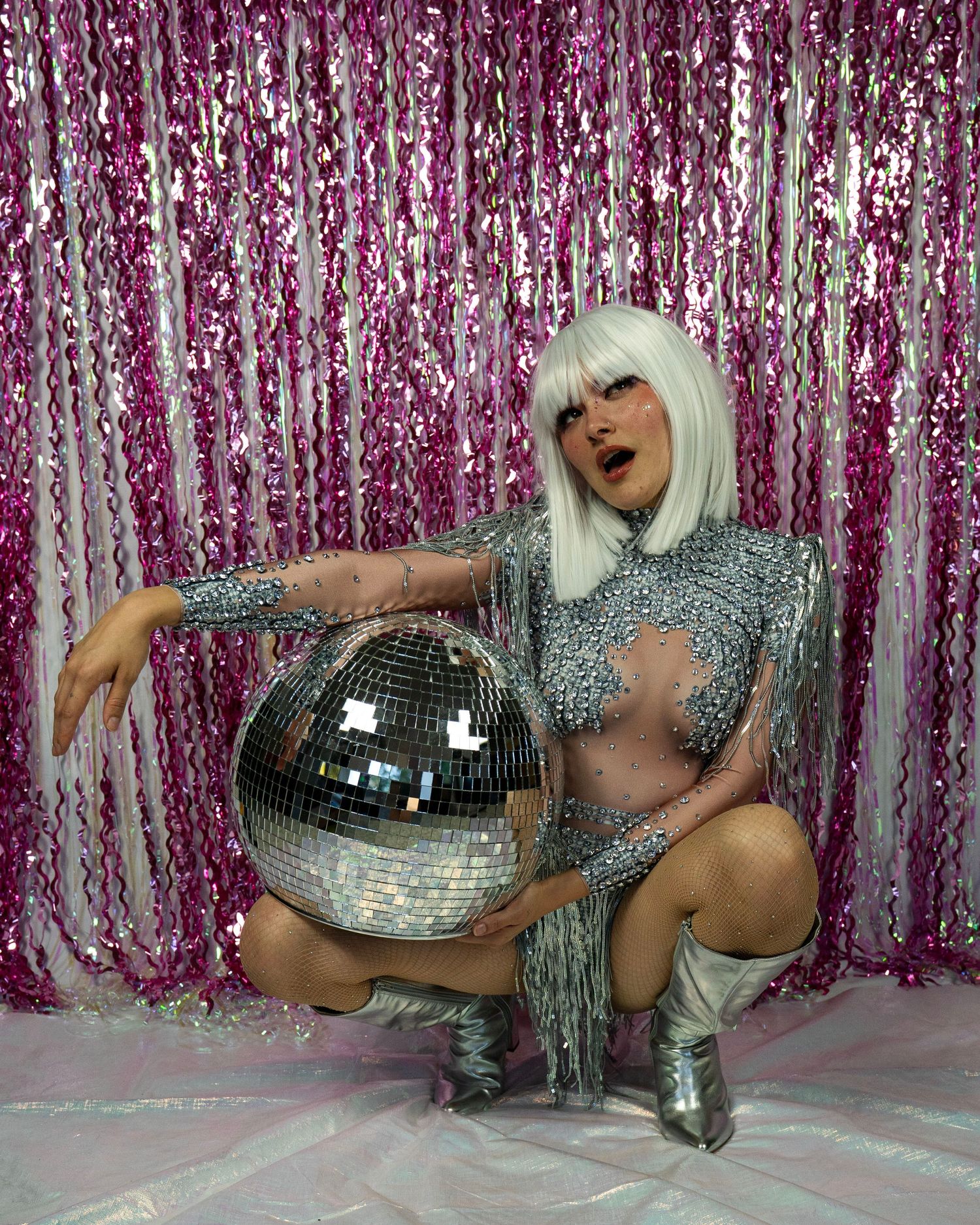 Performer in silver crystal bodysuit poses with mirror disco ball against shimmering pink backdrop.