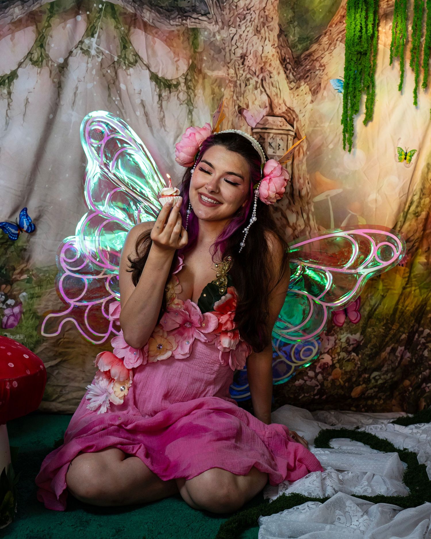 Smiling woman in pink fairy dress with glowing wings holds a small candle in enchanted forest set. Fantasy Photoshoot