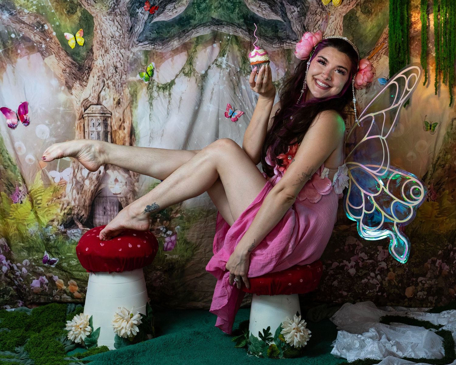Playful woman in pink fairy dress with glowing wings poses on red mushroom in magical enchanted forest backdrop. Fantasy Photoshoot