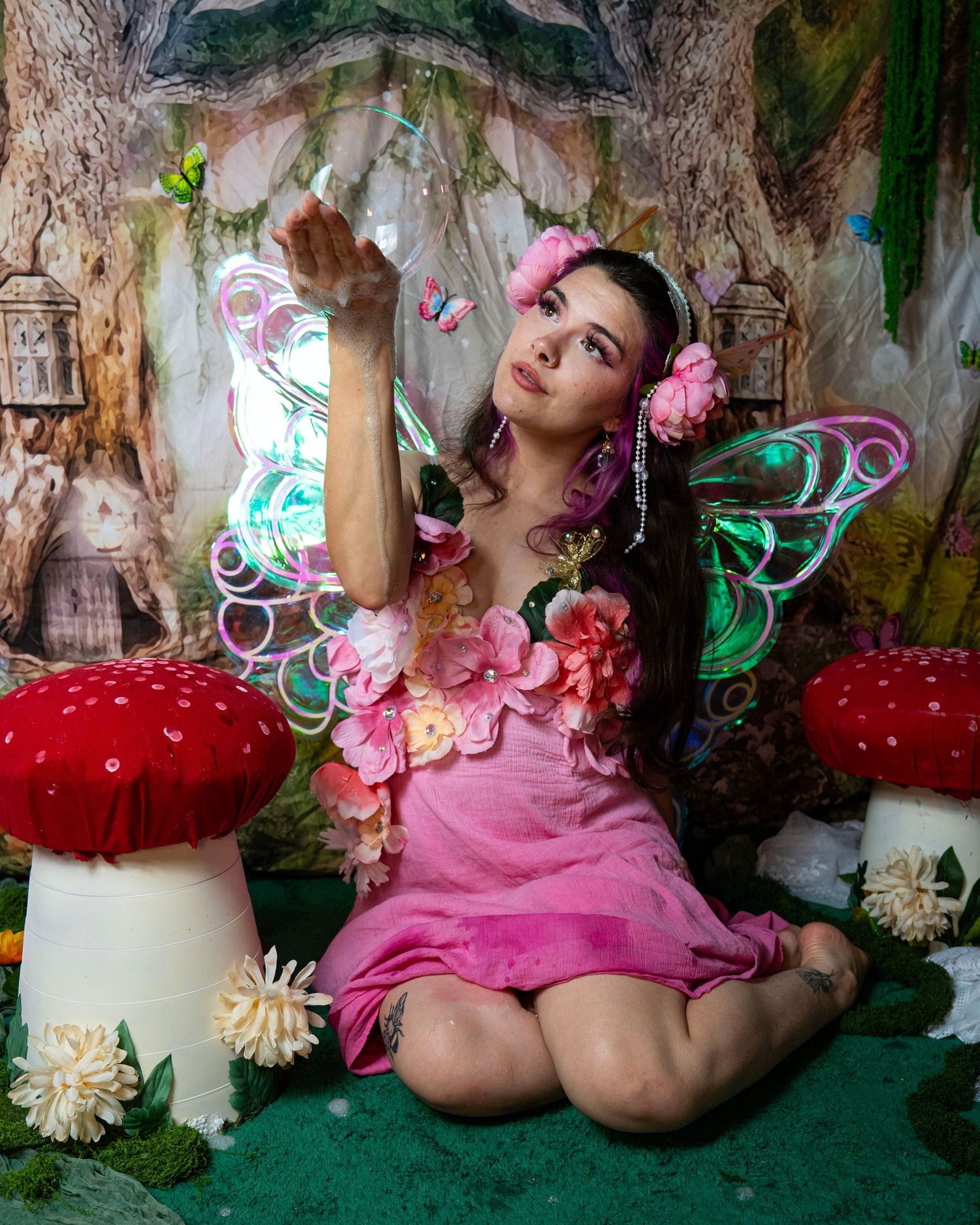 Fairy cosplay model in pink dress with colorful wings poses among giant red mushrooms in magical forest backdrop. Fantasy Photoshoot