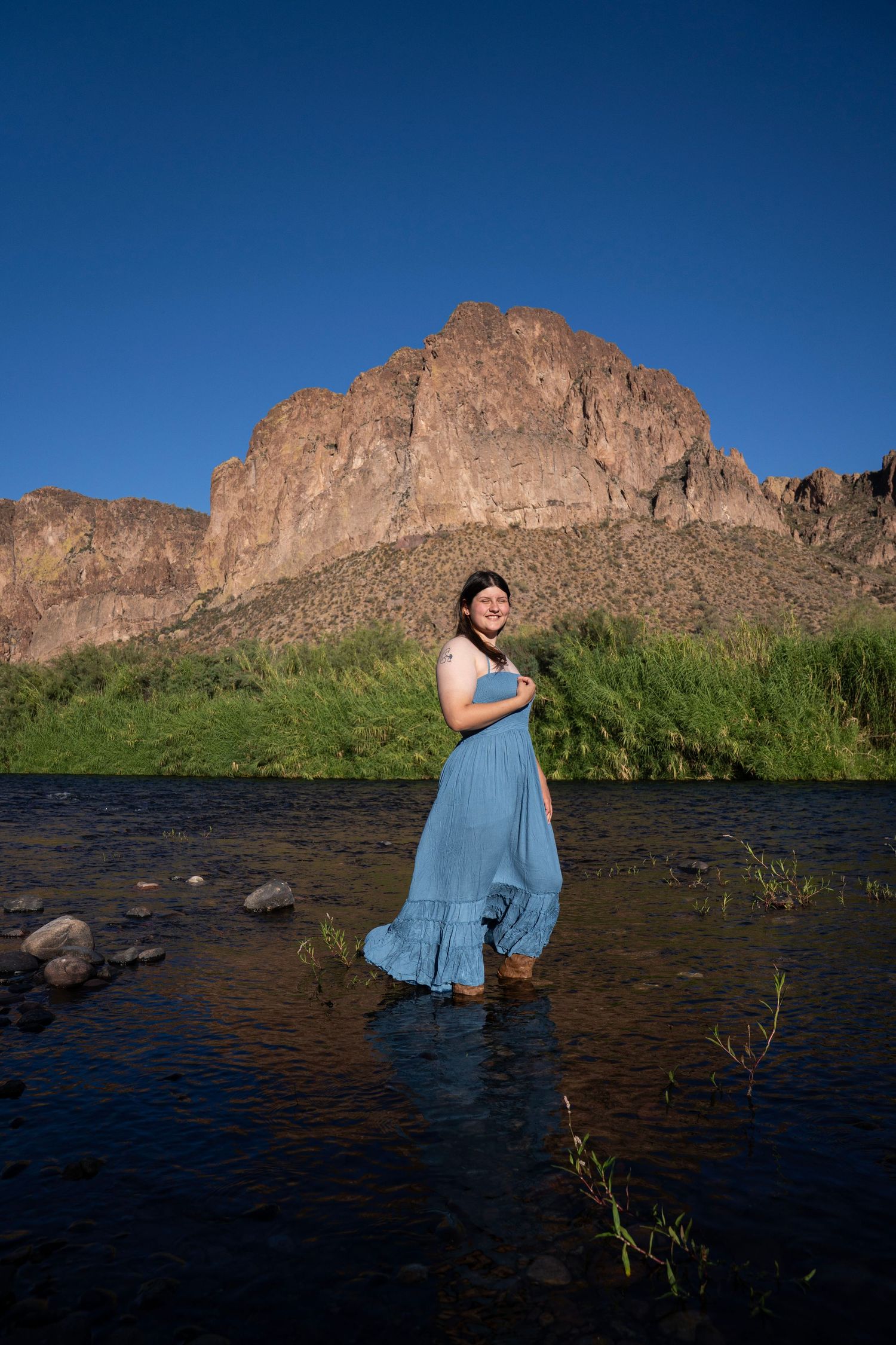 Pregnant woman in blue dress standing in a shallow river with rocky mountain and green trees in background.