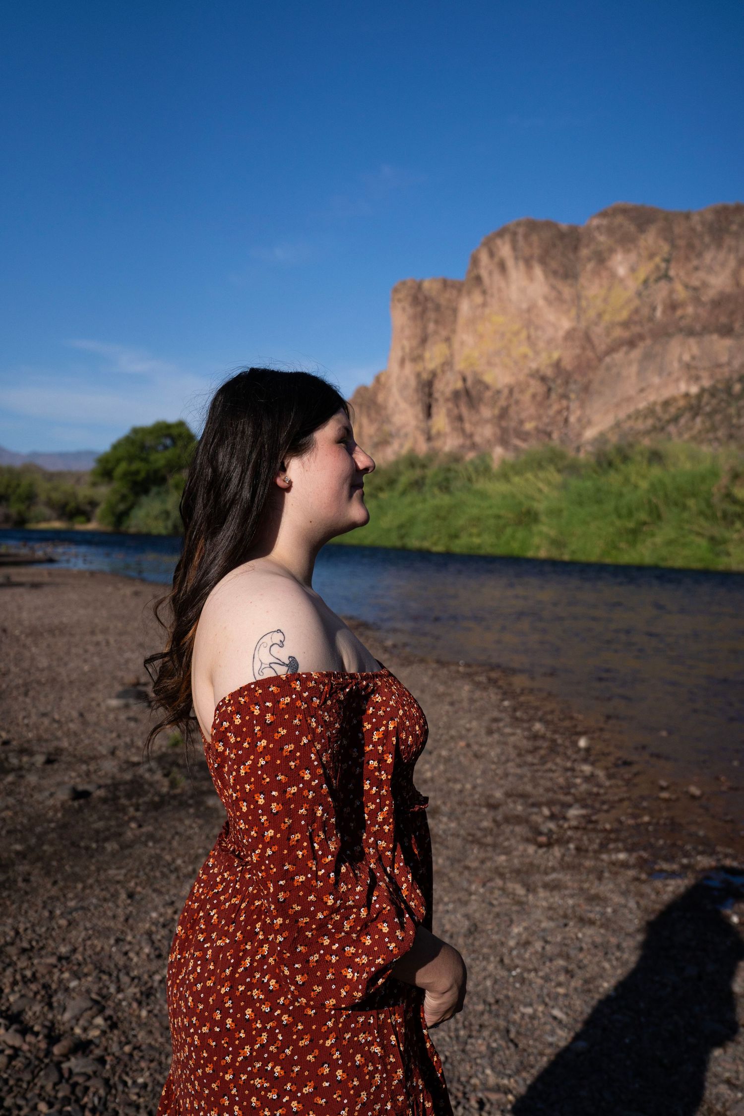 Woman in red floral off-shoulder dress stands by a river with rocky cliffs and blue sky backdrop.