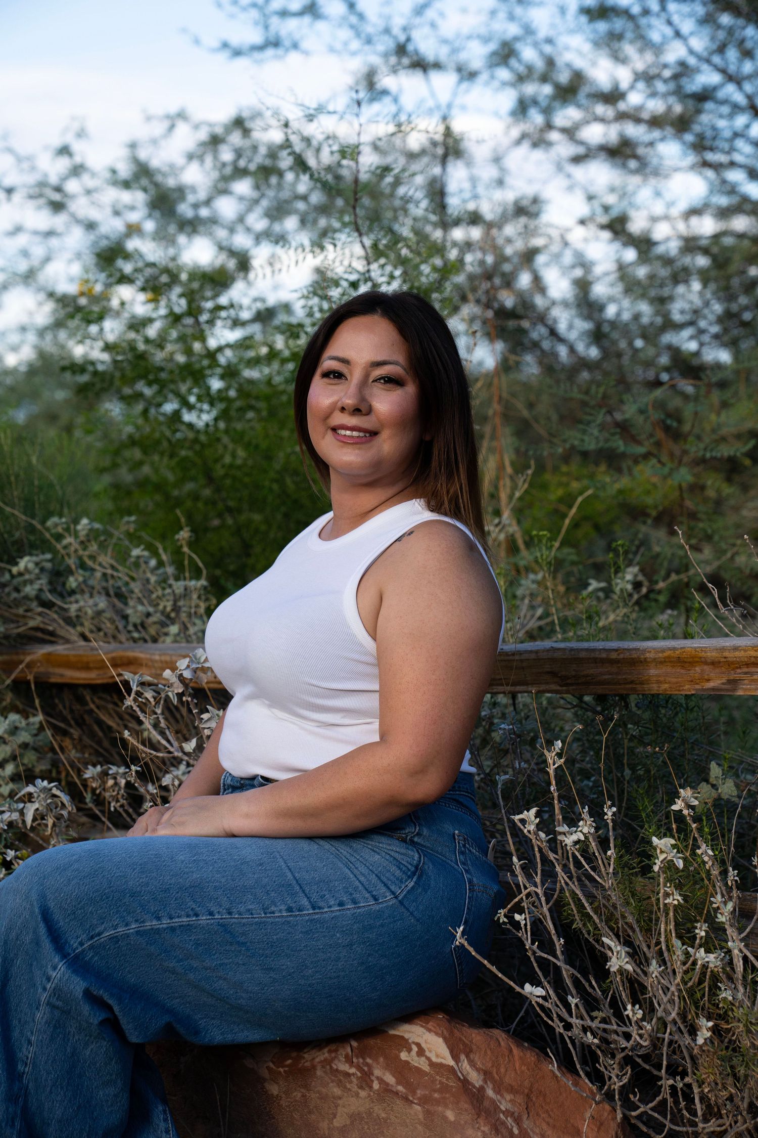 A smiling woman in a white tank top and jeans sits on a rock outdoors, surrounded by lush green desert vegetation.