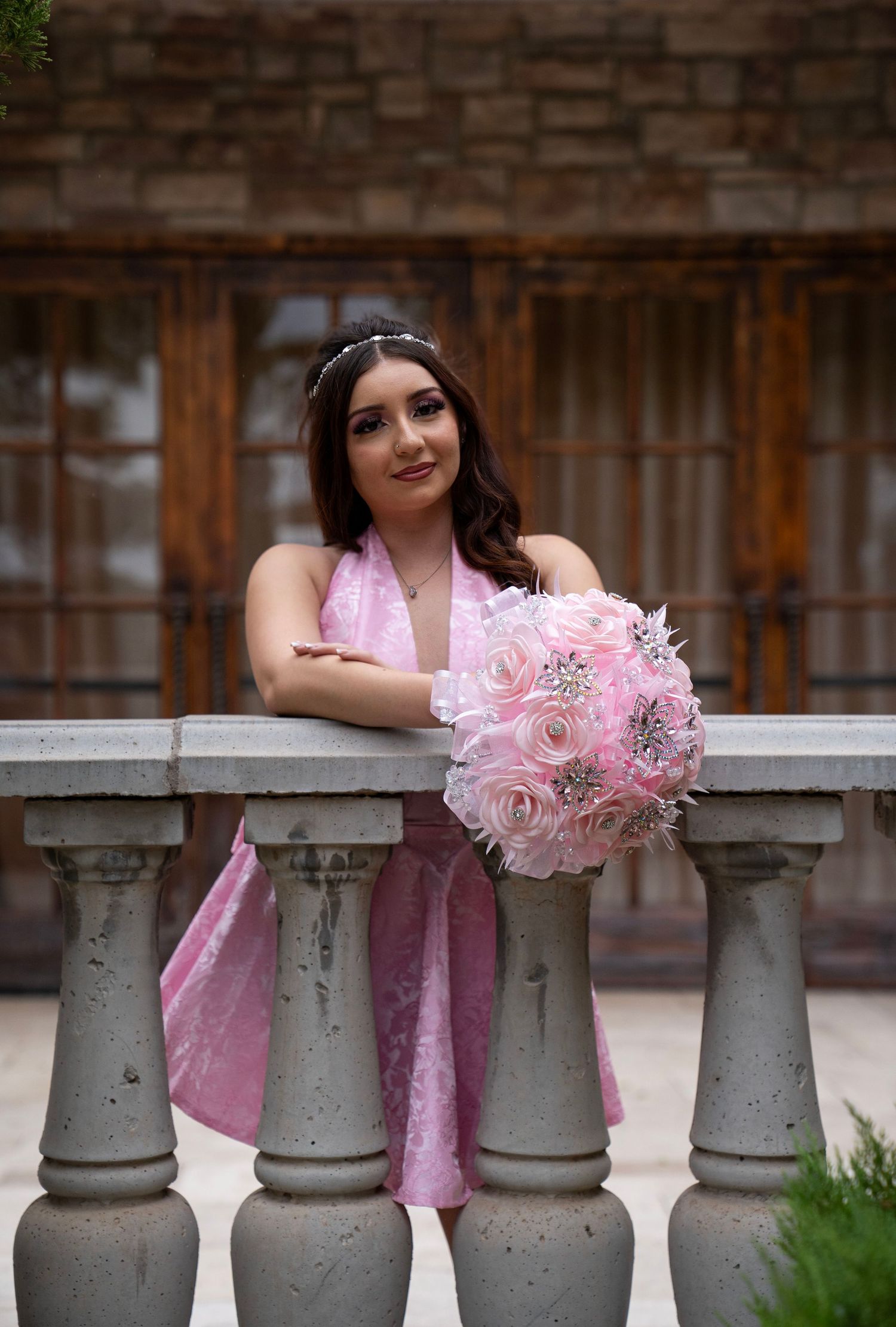 Elegant young woman in pink dress leaning on stone balustrade holding pink brooch bouquet.