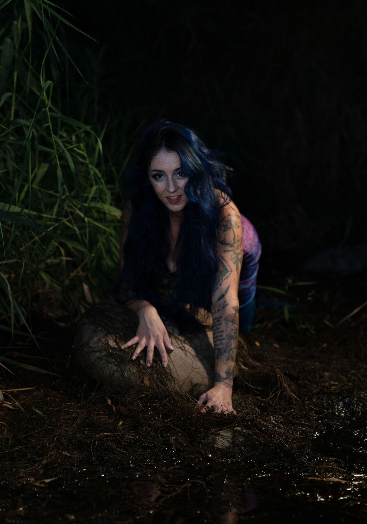 Tattooed woman with blue-black hair crouching on forest ground at night in dark atmospheric photo.