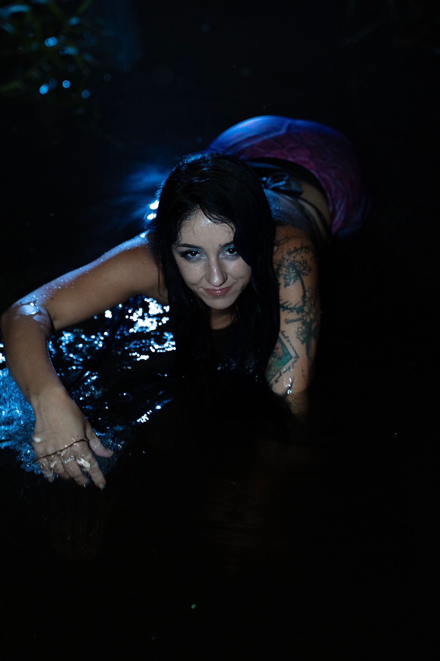 Tattooed woman in mermaid costume crawling through dark illuminated water at night.