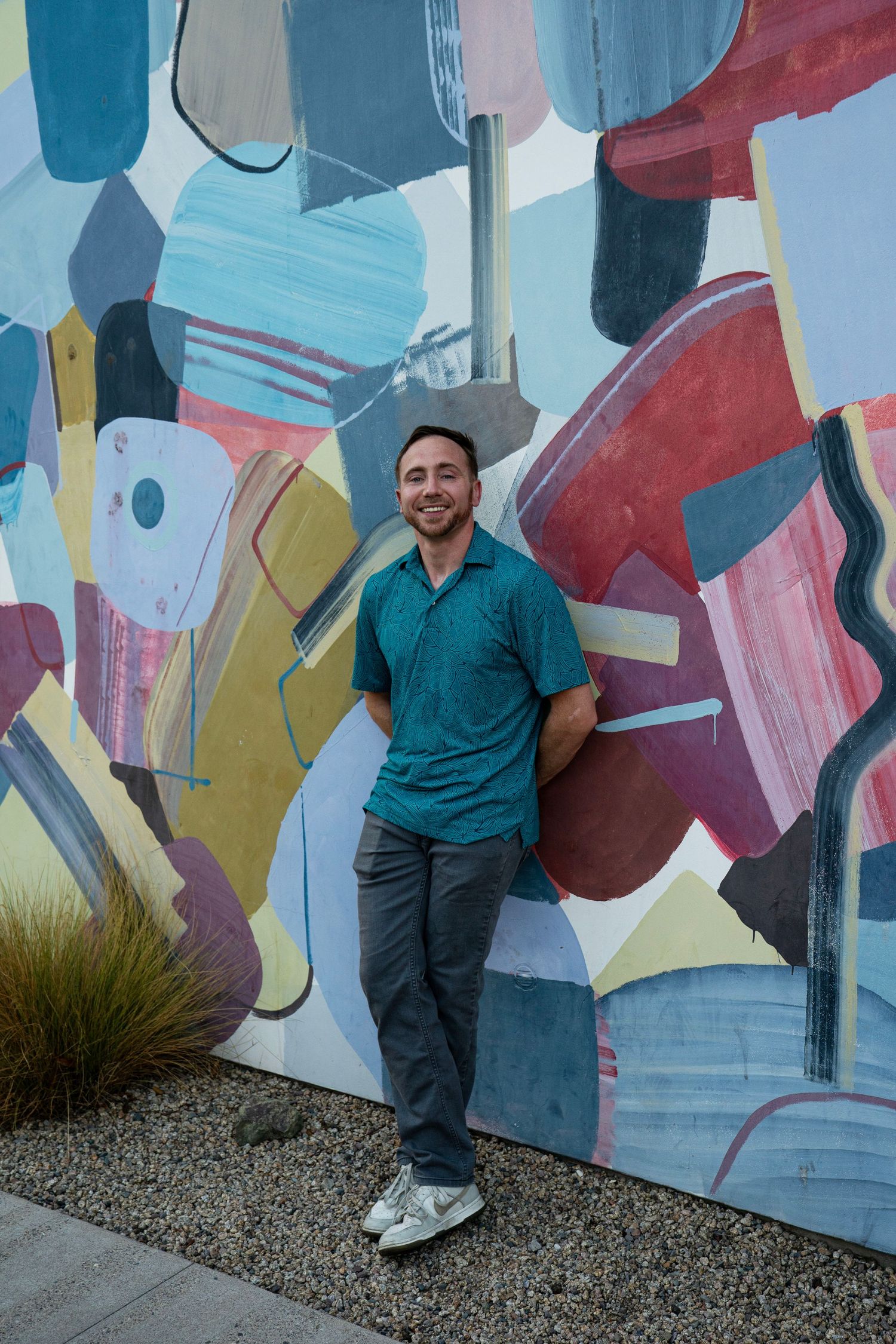 Smiling man in teal polo shirt leaning against a colorful abstract mural wall outdoors.