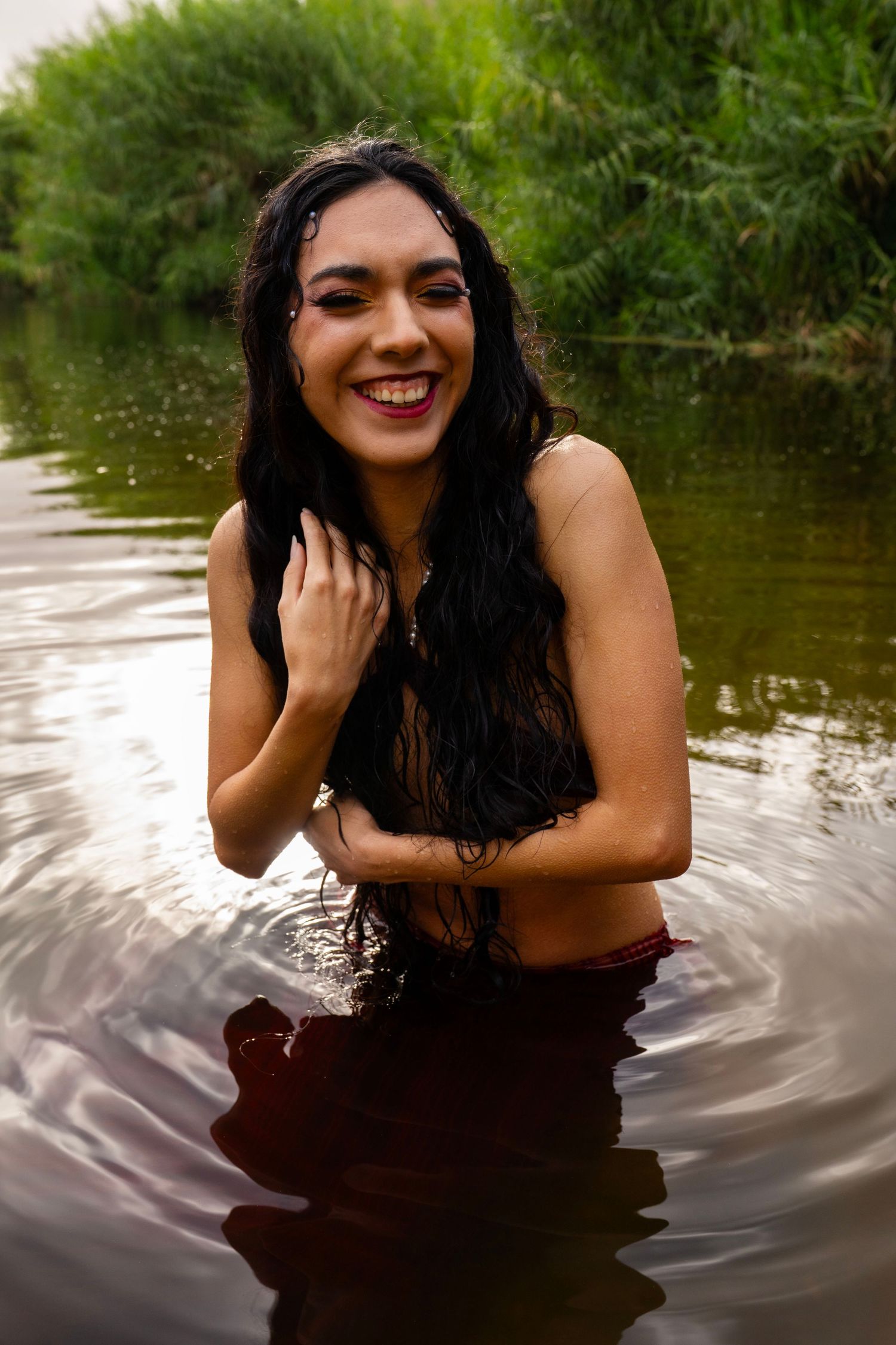 A smiling woman with long wet dark hair stands in a river wearing a red skirt in a joyful outdoor photoshoot.