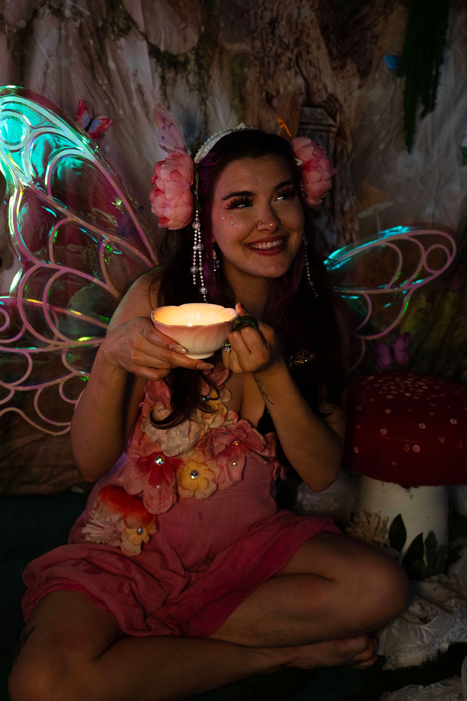 Cheerful woman in floral fairy costume holds glowing candle cup while seated near red mushroom prop. Fantasy Photoshoot