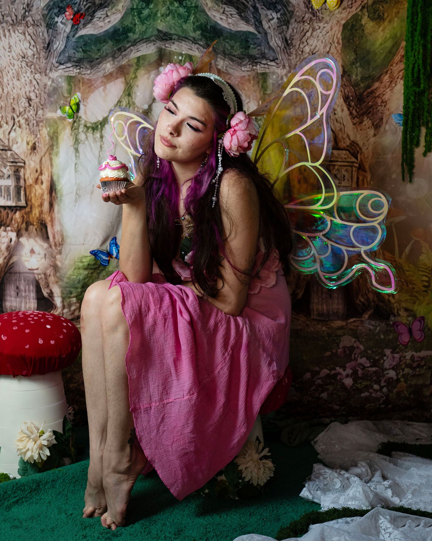 Fairy-costumed woman with purple hair and glowing wings closes eyes while holding cupcake in enchanted forest backdrop. Fantasy Photoshoot