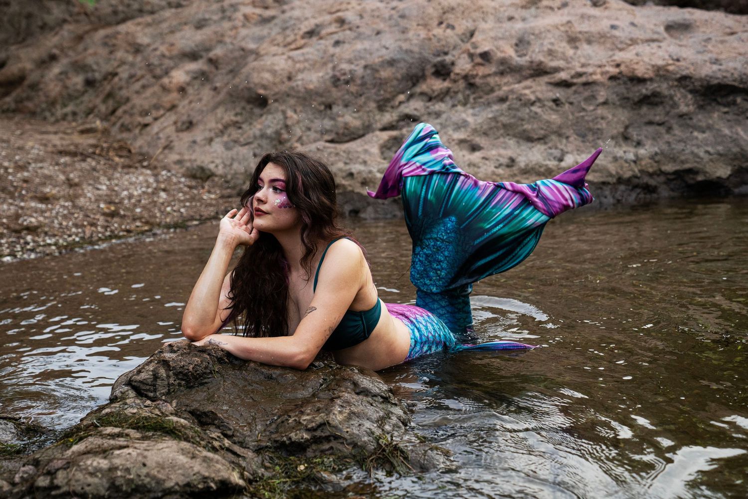 A brunette woman in a mermaid costume with colorful tail poses on rocks in shallow water. Fantasy Photoshoot