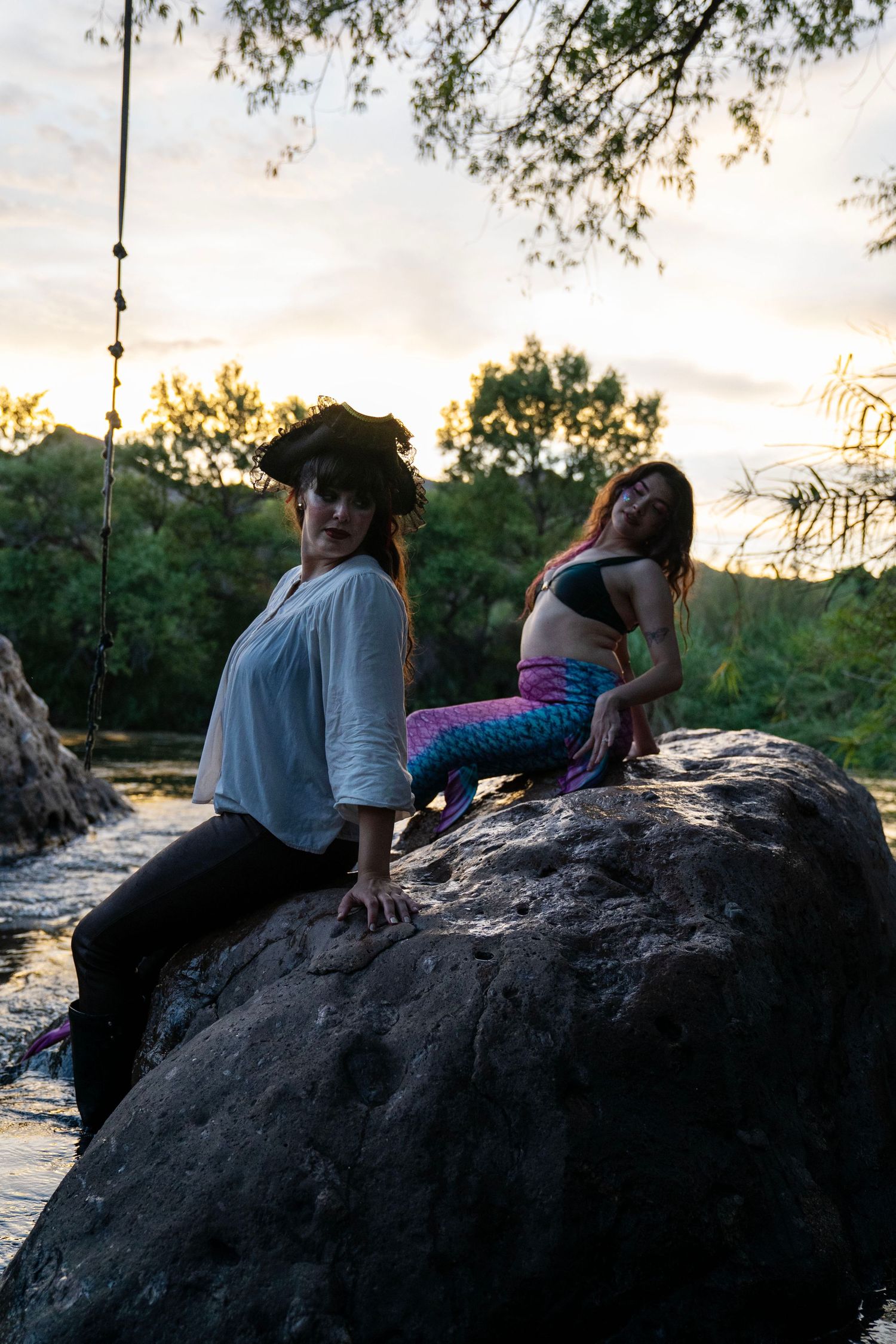Pirate and mermaid costumed women pose together on large rock at river during sunset. Mermaid photoshoot, fantasy photoshoot.
