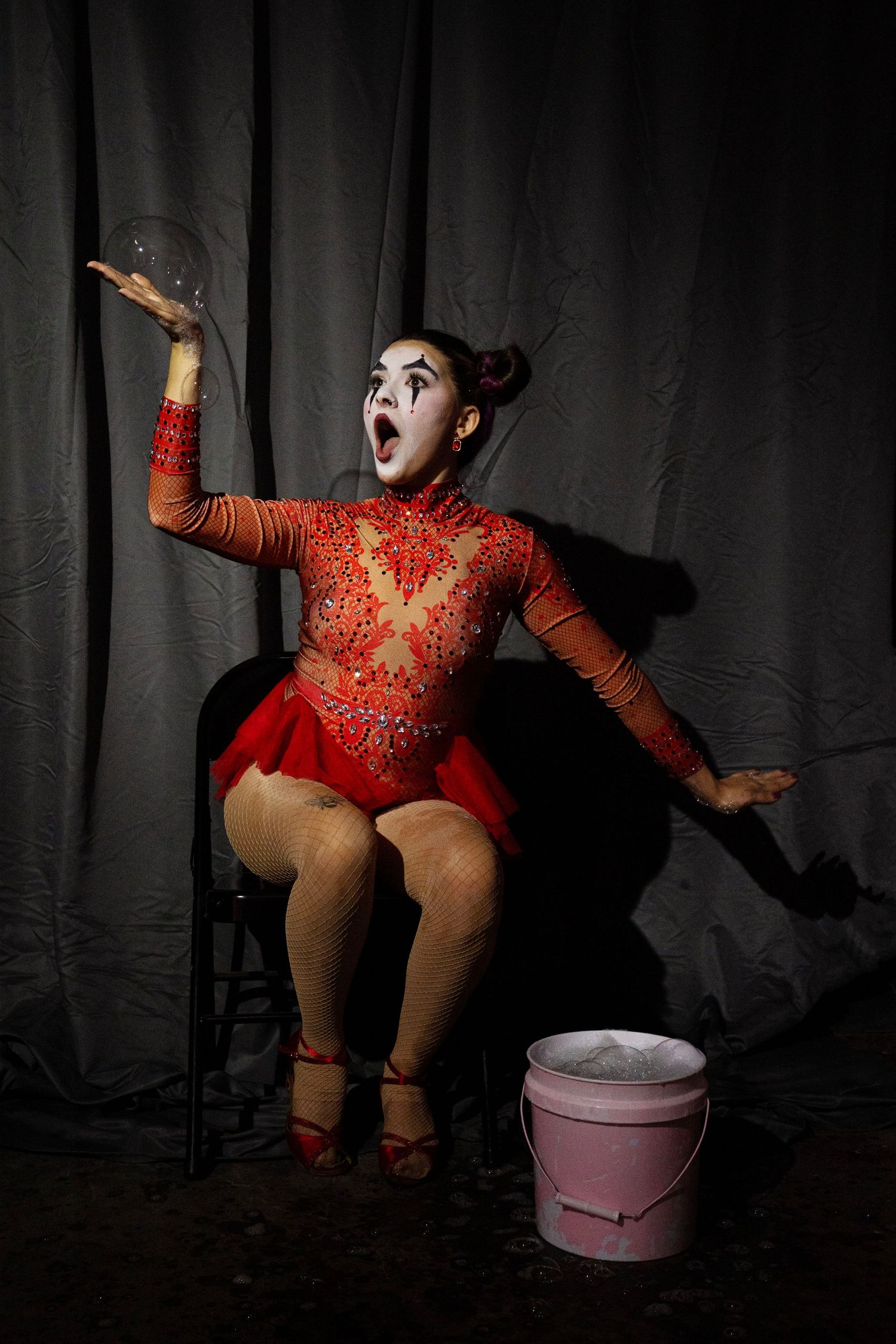 A circus clown in red sequined dress reacts with surprise to a soap bubble beside a pink bucket.