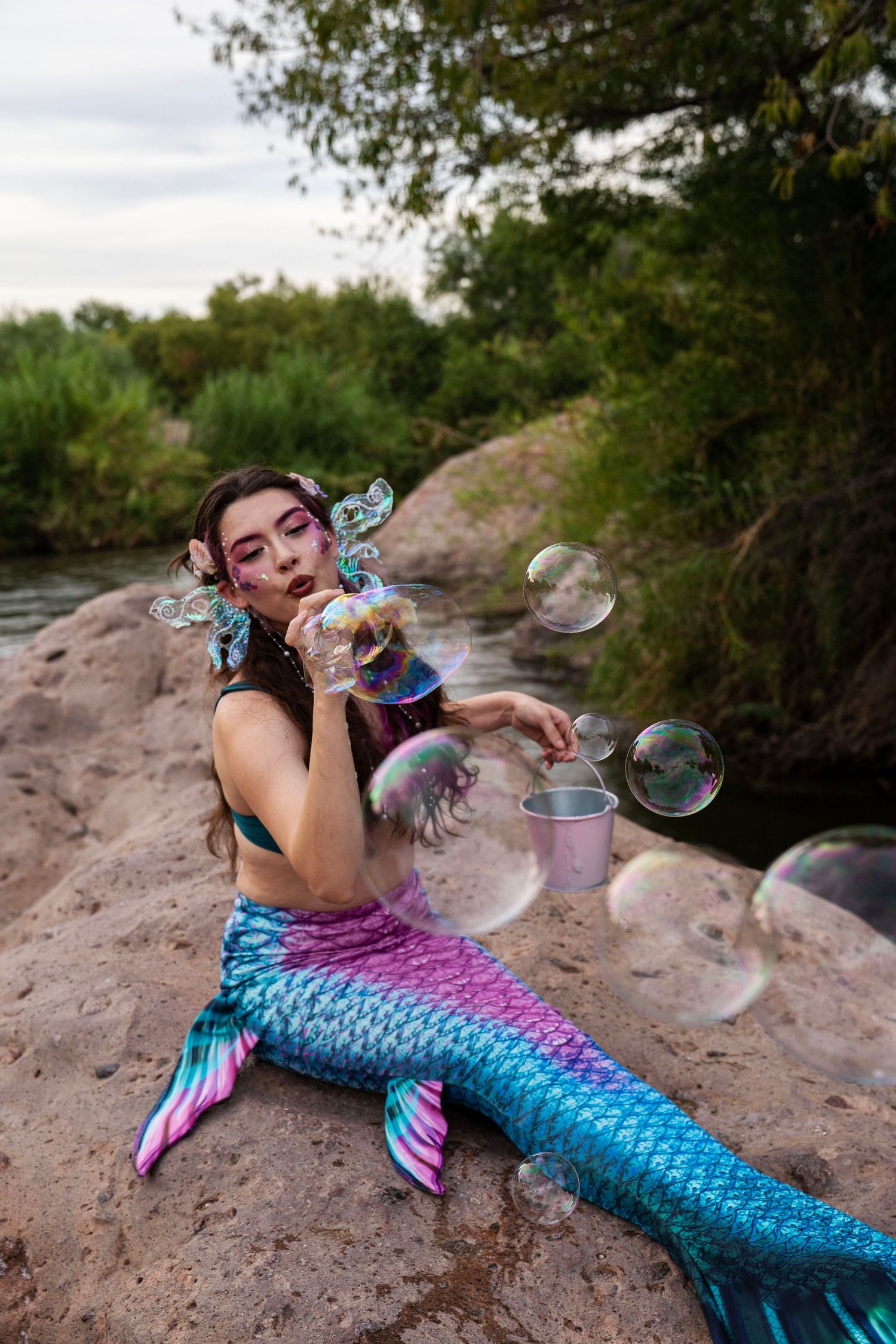 Woman in blue and pink mermaid tail blowing soap bubbles surrounded by floating bubbles on riverbank. Fantasy Photoshoot