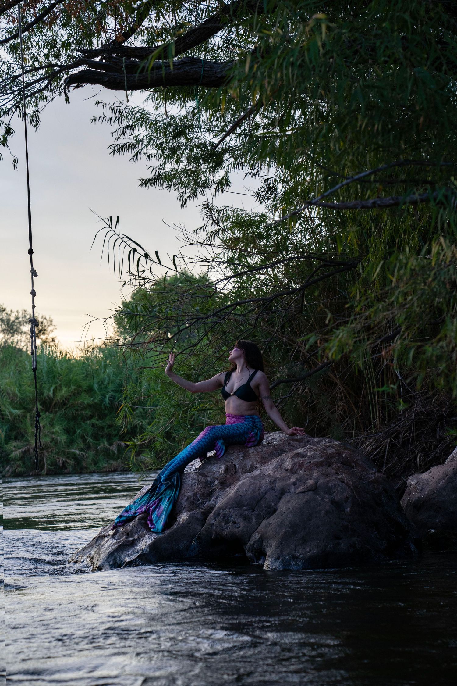 A woman in a mermaid costume sits on rocks by a river at dusk, reaching toward overhanging tree branches. Fantasy Photoshoot