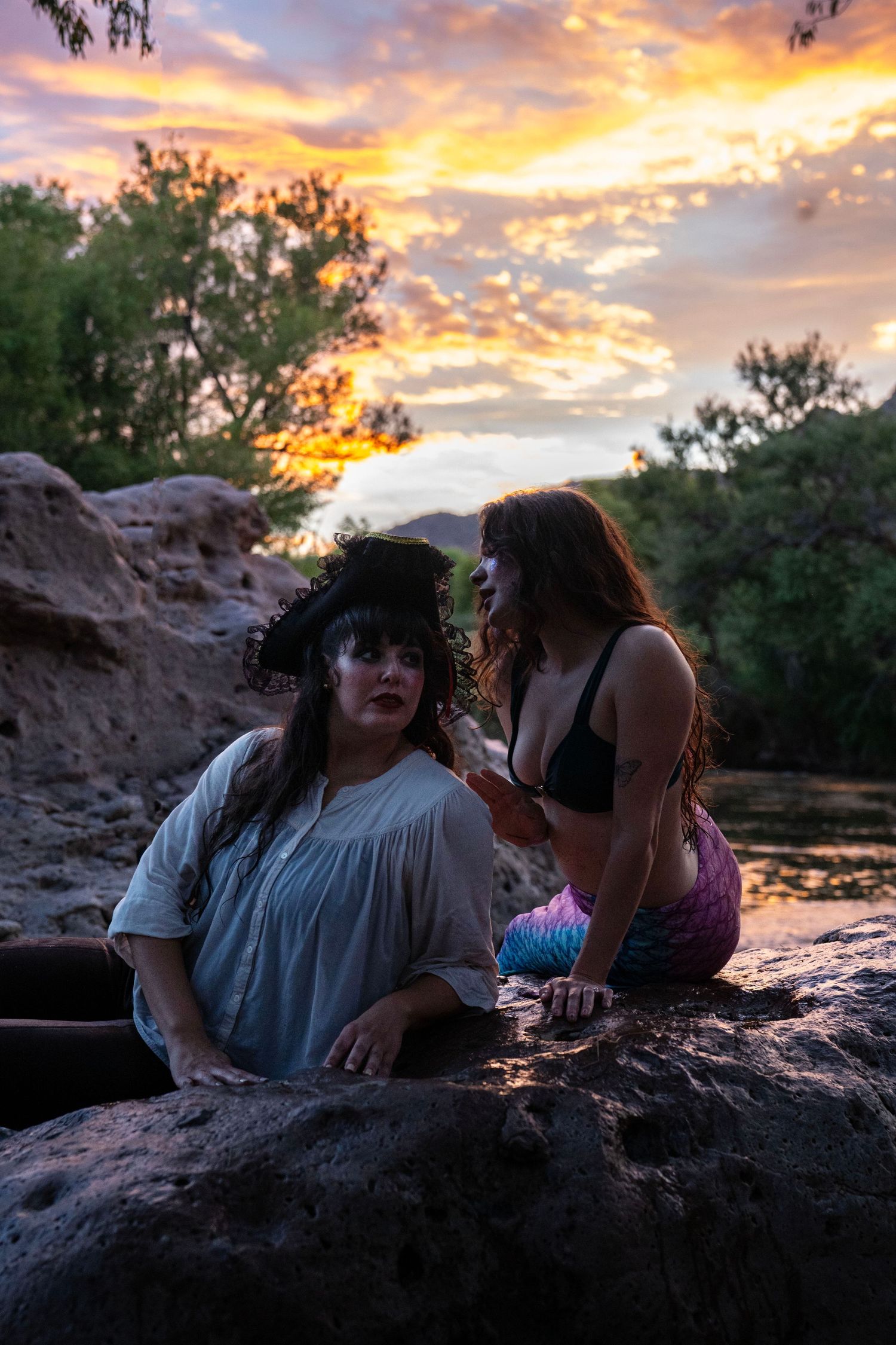 A pirate and mermaid cosplay duo pose dramatically on rocks by a river under a vivid sunset sky. Mermaid photoshoot, fantasy photoshoot.
