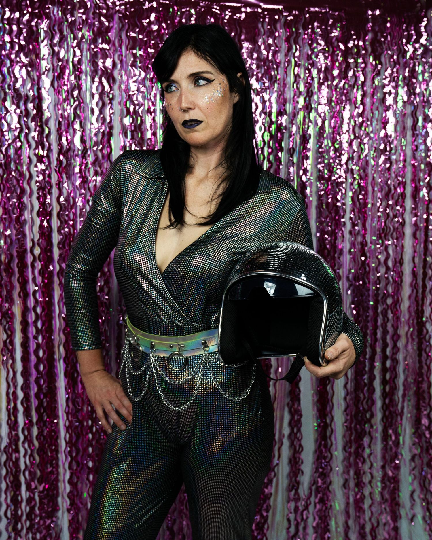 Woman in holographic dark jumpsuit holds black motorcycle helmet against sparkling pink tinsel backdrop with chain belt.