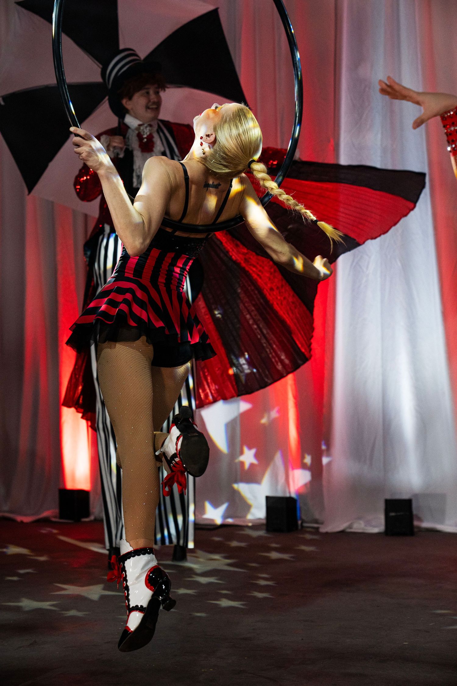 Blonde aerial performer in red and black costume holds hoop with butterfly wings on stage.