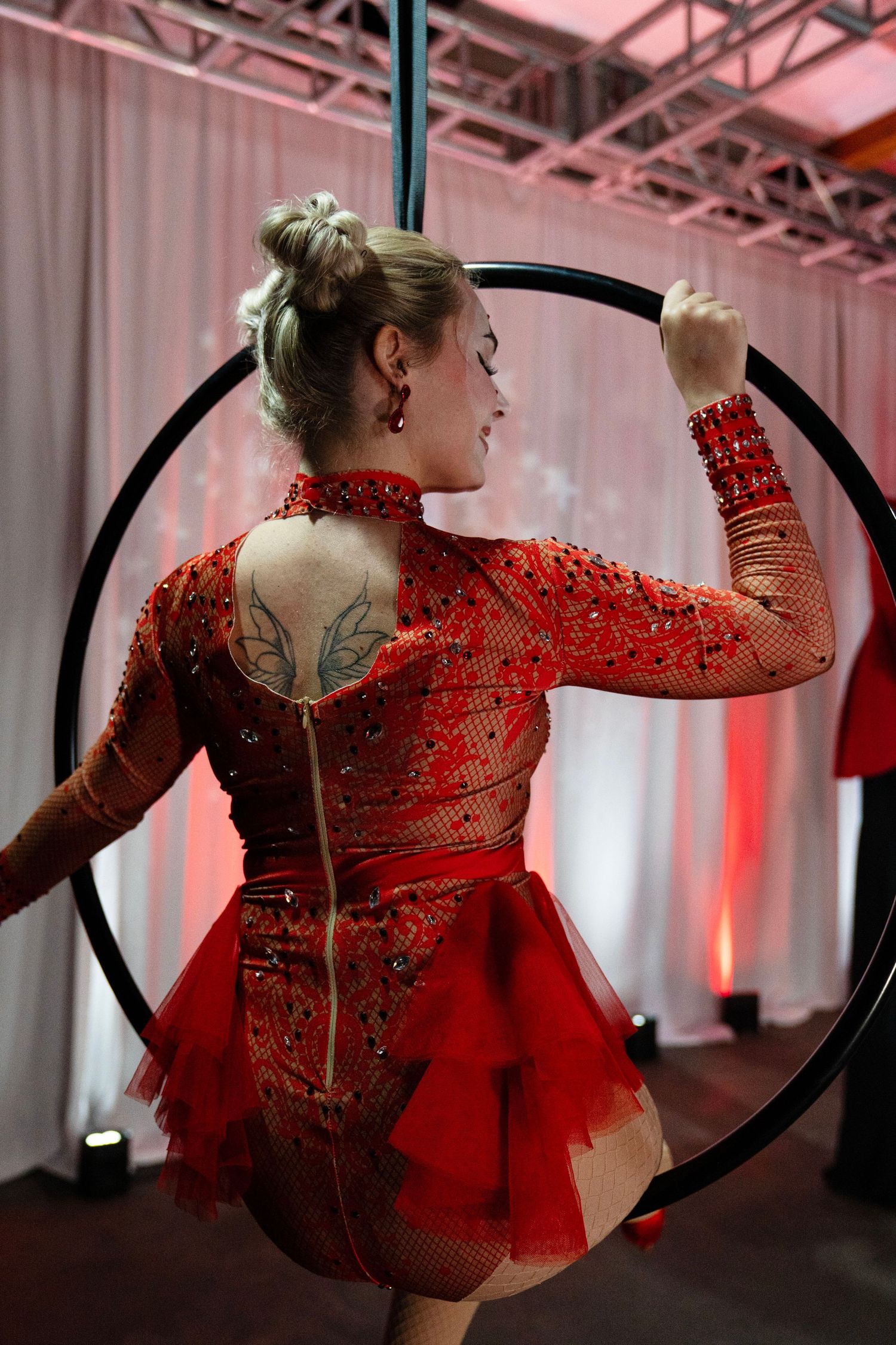 Back view of blonde aerialist in red sequin dress with wing tattoo on lyra.