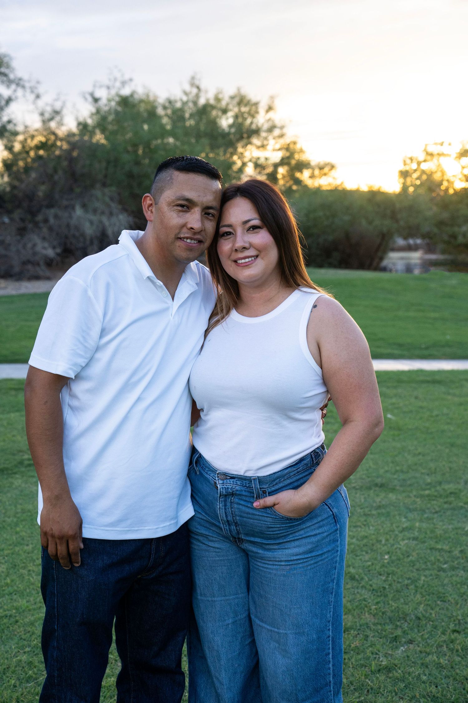 A couple in matching white tops and jeans smiling together outdoors at sunset in a green park.
