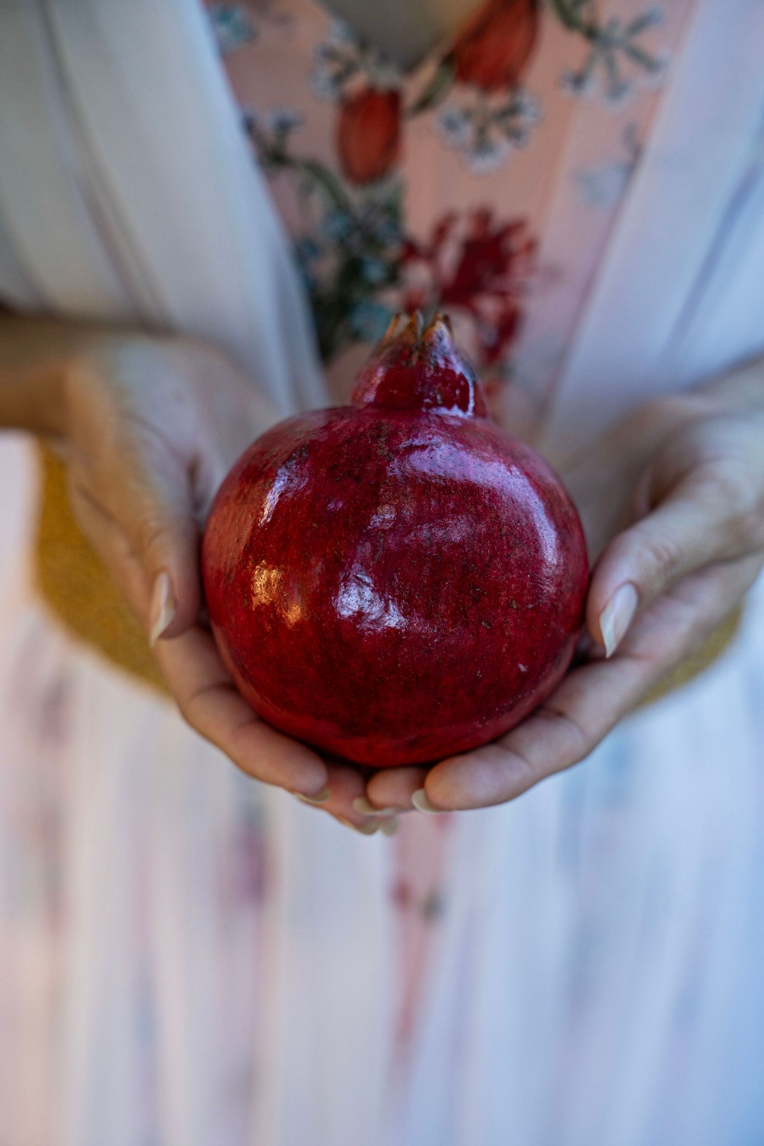 Woman in a floral dress gently holds a ripe red pomegranate in both open hands, showcasing its glossy, deep crimson skin.