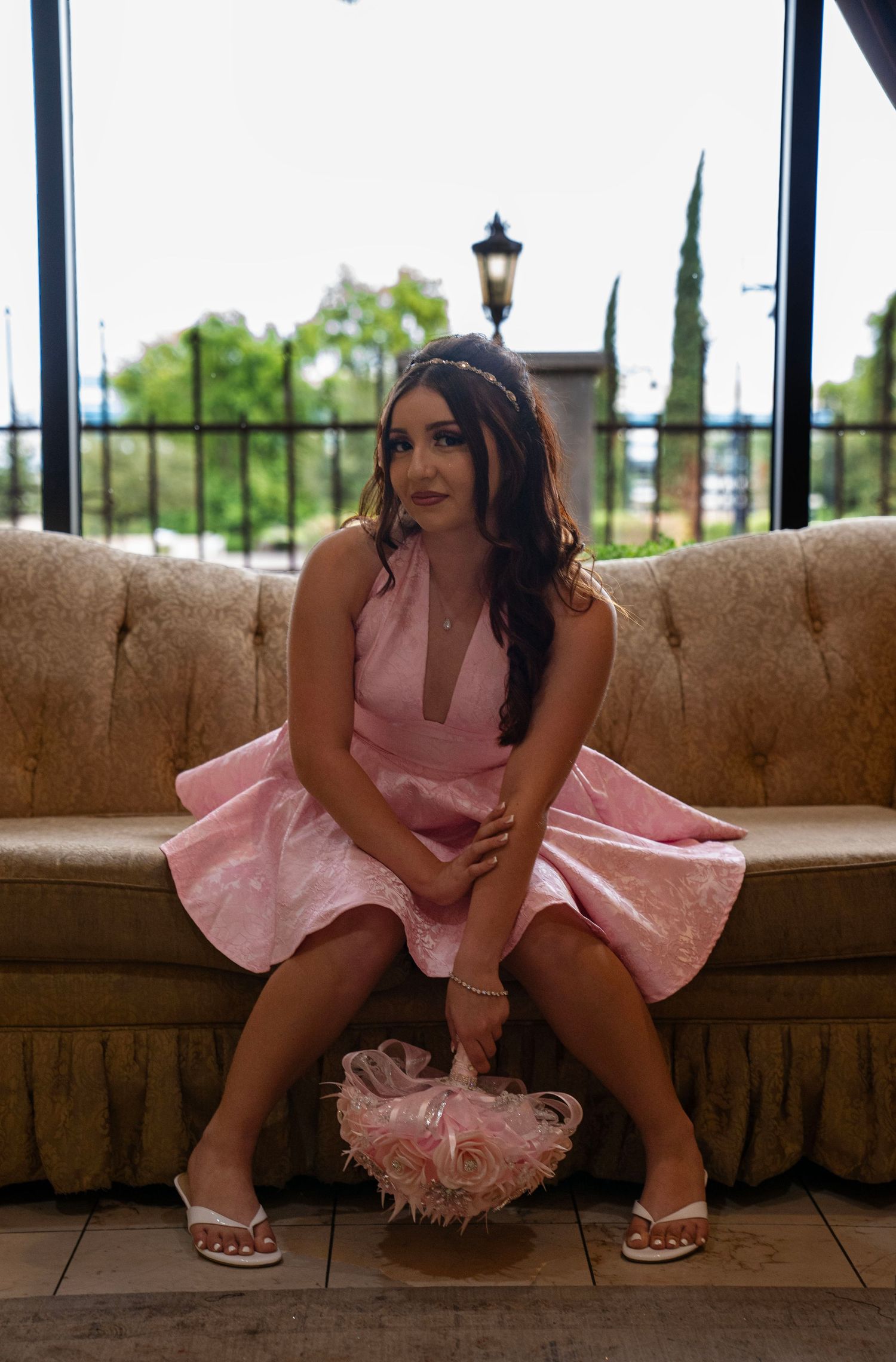 Young woman in pink dress sitting on tufted sofa holding floral accessory in elegant venue.