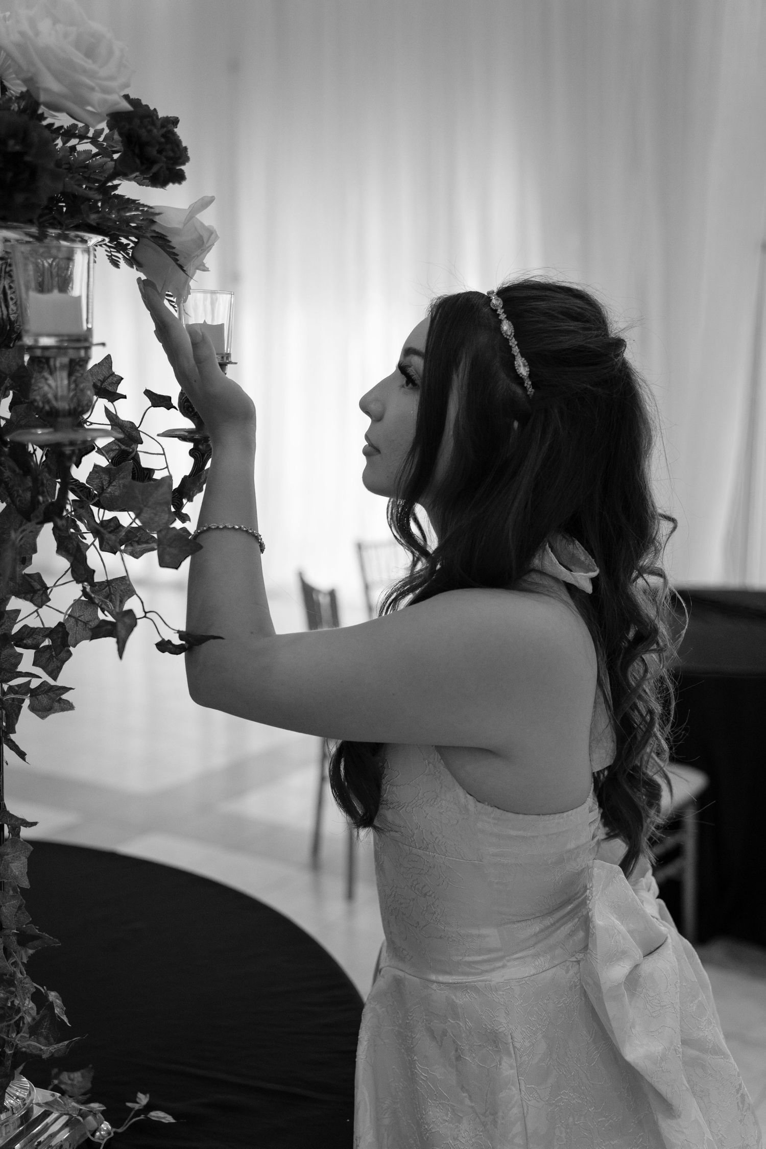 Black and white photo of a bride in a strapless gown arranging floral decorations at her wedding reception.