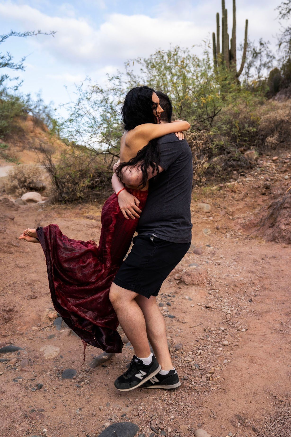 Two people embracing on a desert trail with saguaro cactus and red mermaid dress flowing.