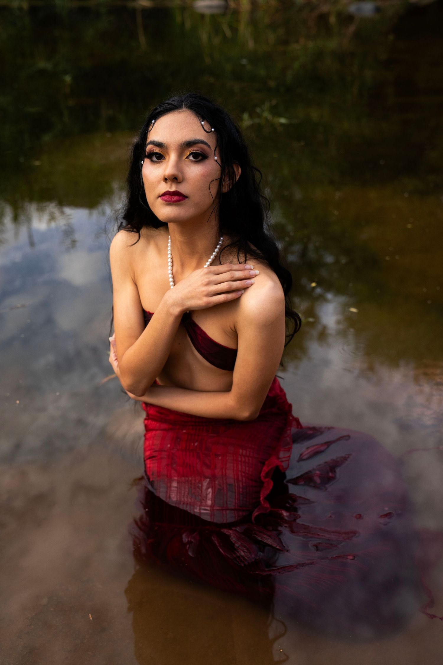 A dark-haired woman wearing a pearl necklace and red skirt kneels in a river, crossing her arms over her chest.