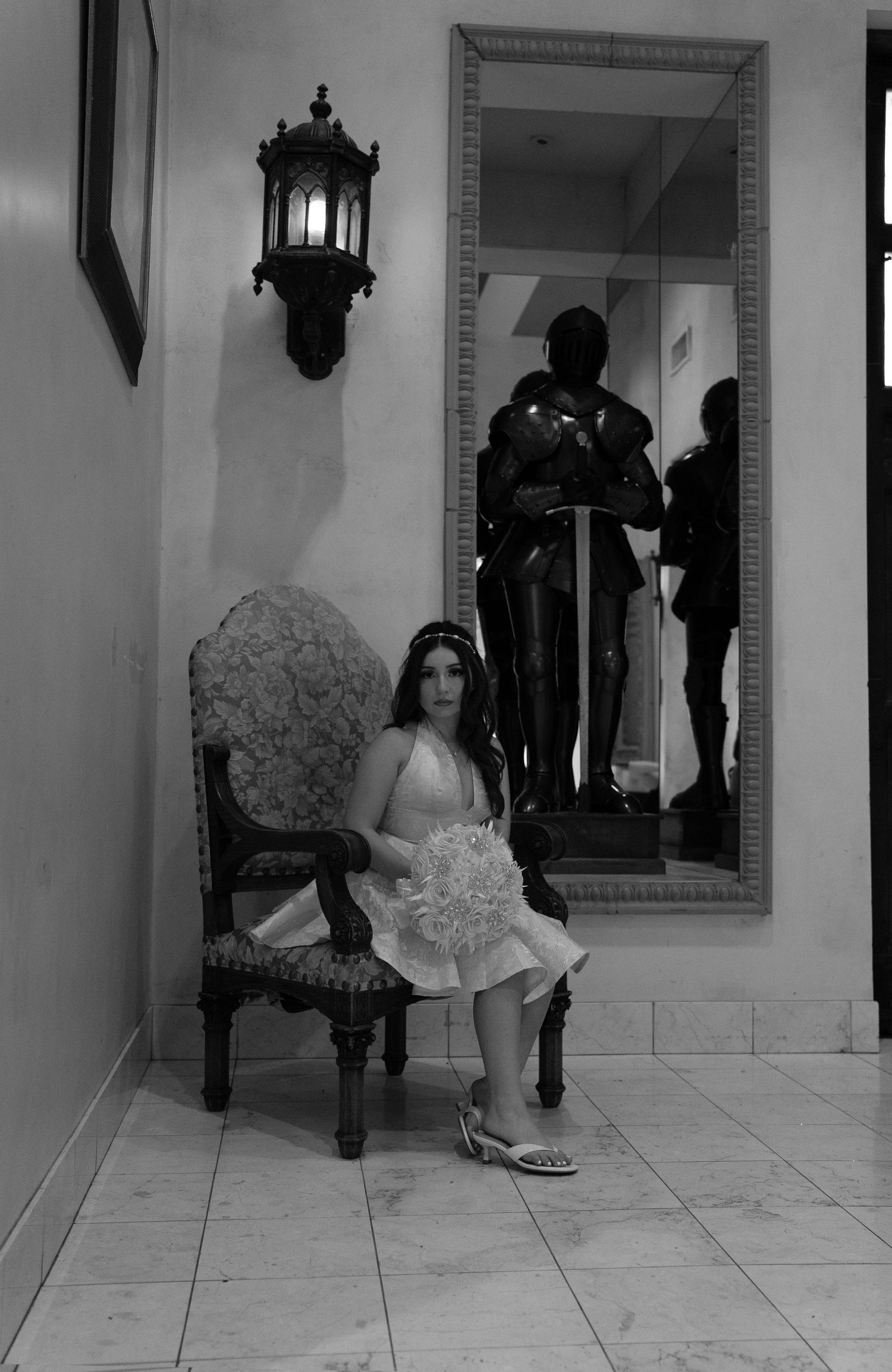 Black and white photo of young woman in white dress seated in ornate chair near mirror reflecting knight armor.