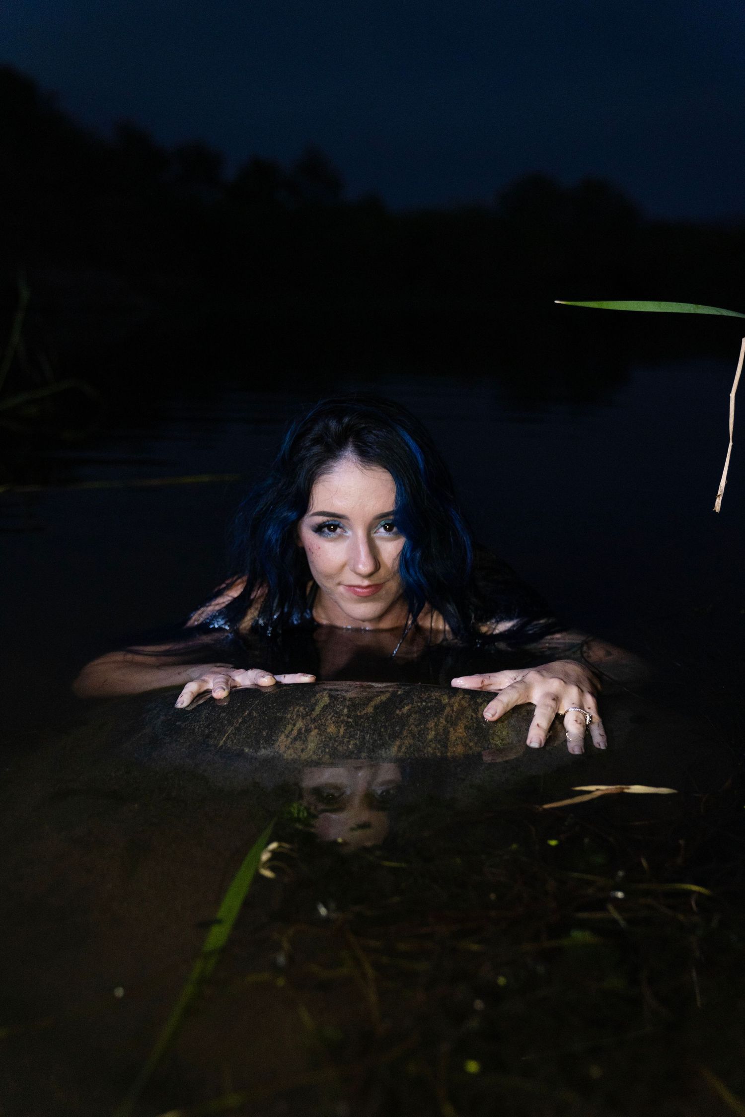 Blue-haired woman with tattoos emerges from dark water gripping a rock at night.