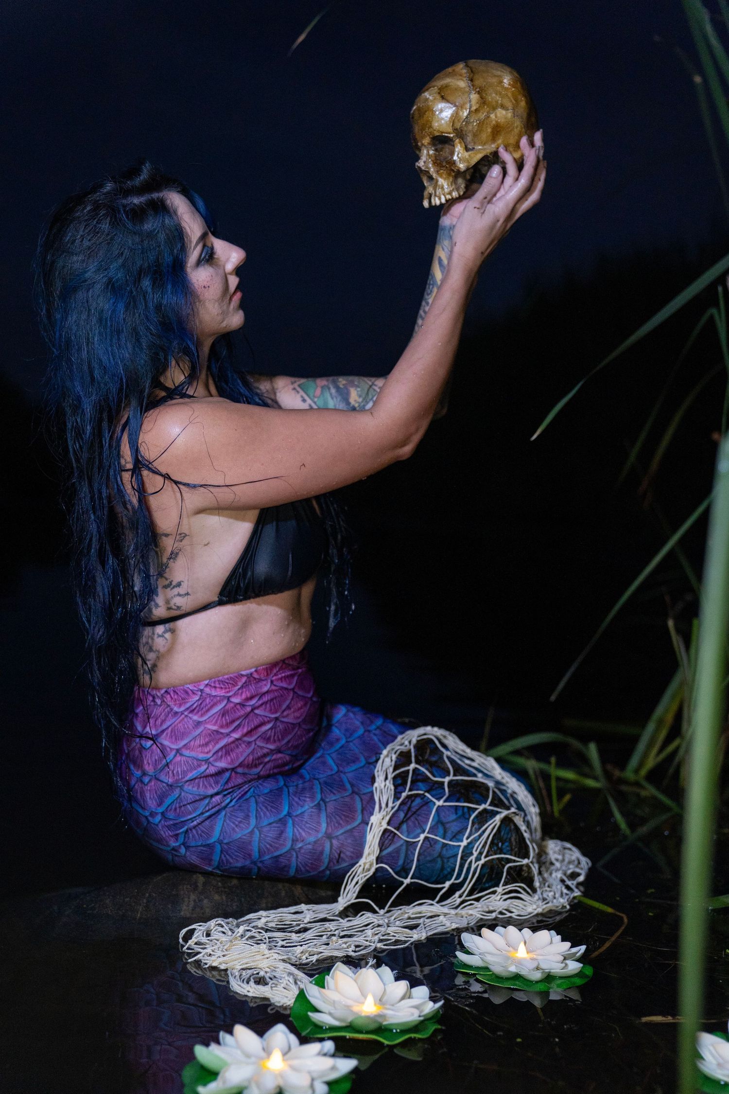 Side view of tattooed mermaid costume model holding skull near lotus flowers at night.
